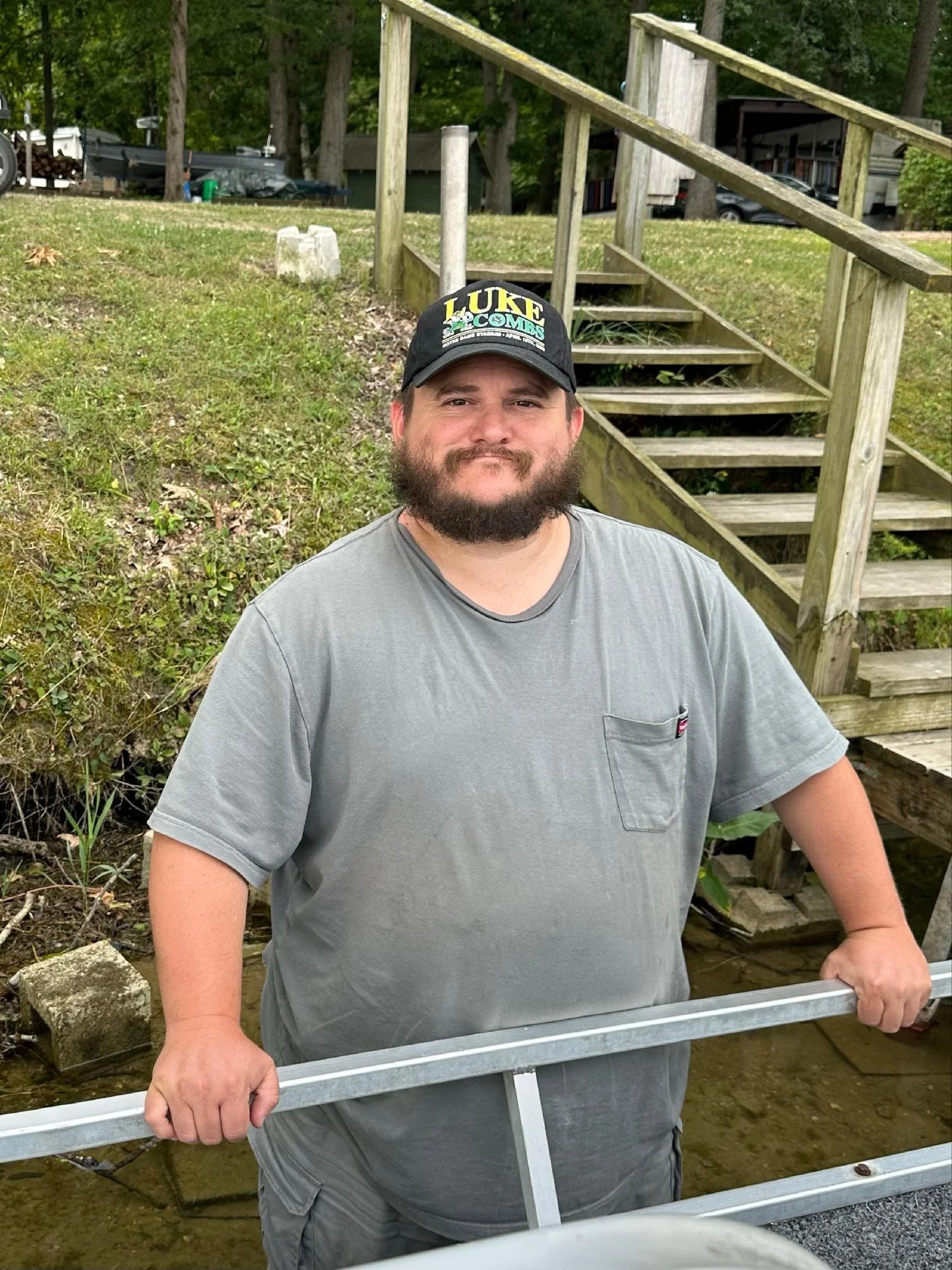 A man with a beard wearing a guide cap and a gray t-shirt, standing outdoors in front of a wooden staircase and greenery, holding a metal frame.