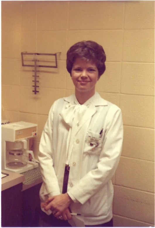 A female healthcare professional in a white coat standing in a medical office with a beige tiled wall, a wall-mounted medical equipment rack, and a coffee maker on the counter.