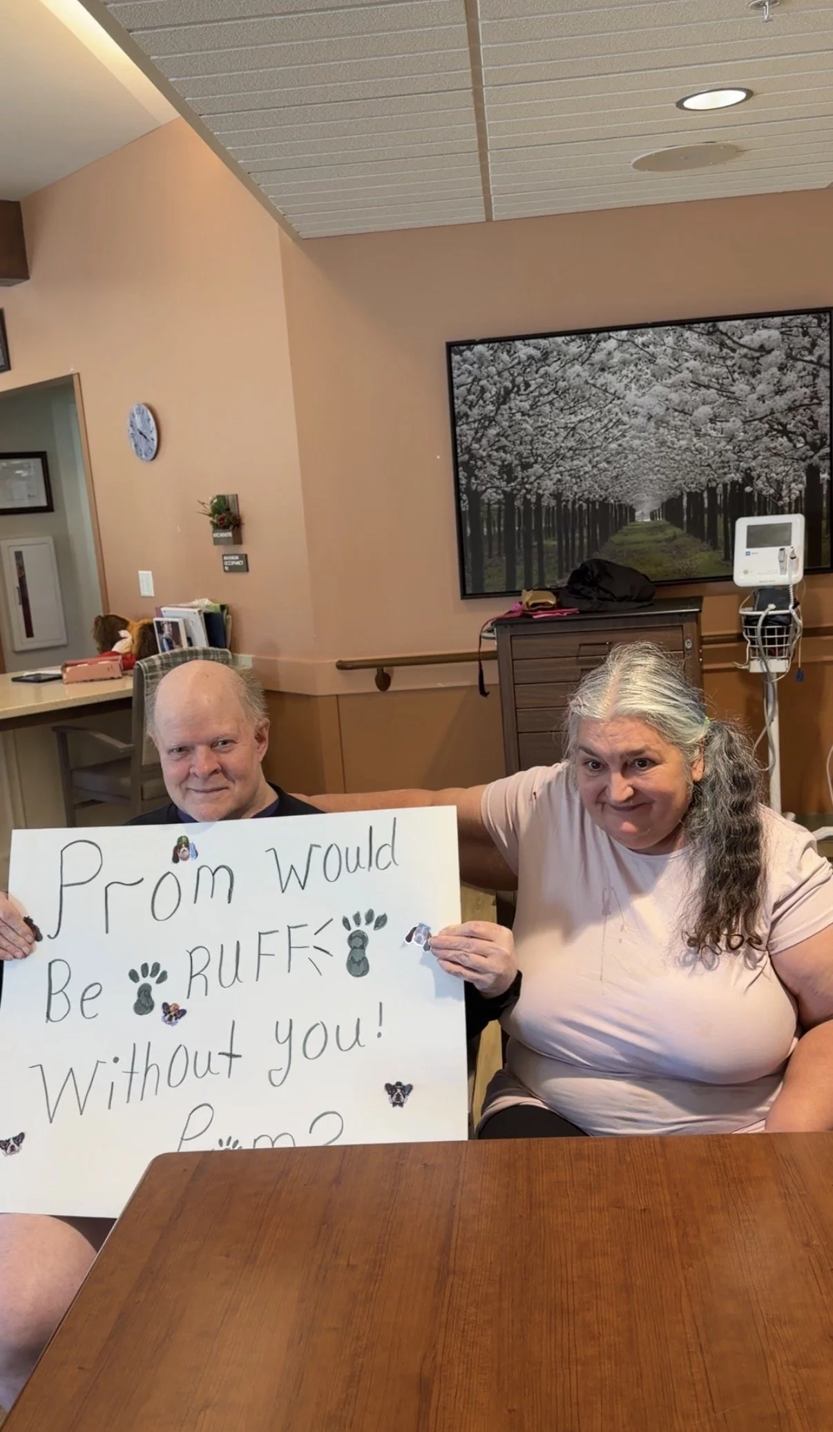 Two women sitting at a table, one holding a sign that reads, "Prom would be RUFF without you!" The woman on the right has long gray hair, and the woman on the left has three-quarter gray hair. Behind them is a wall with a large framed picture of cher