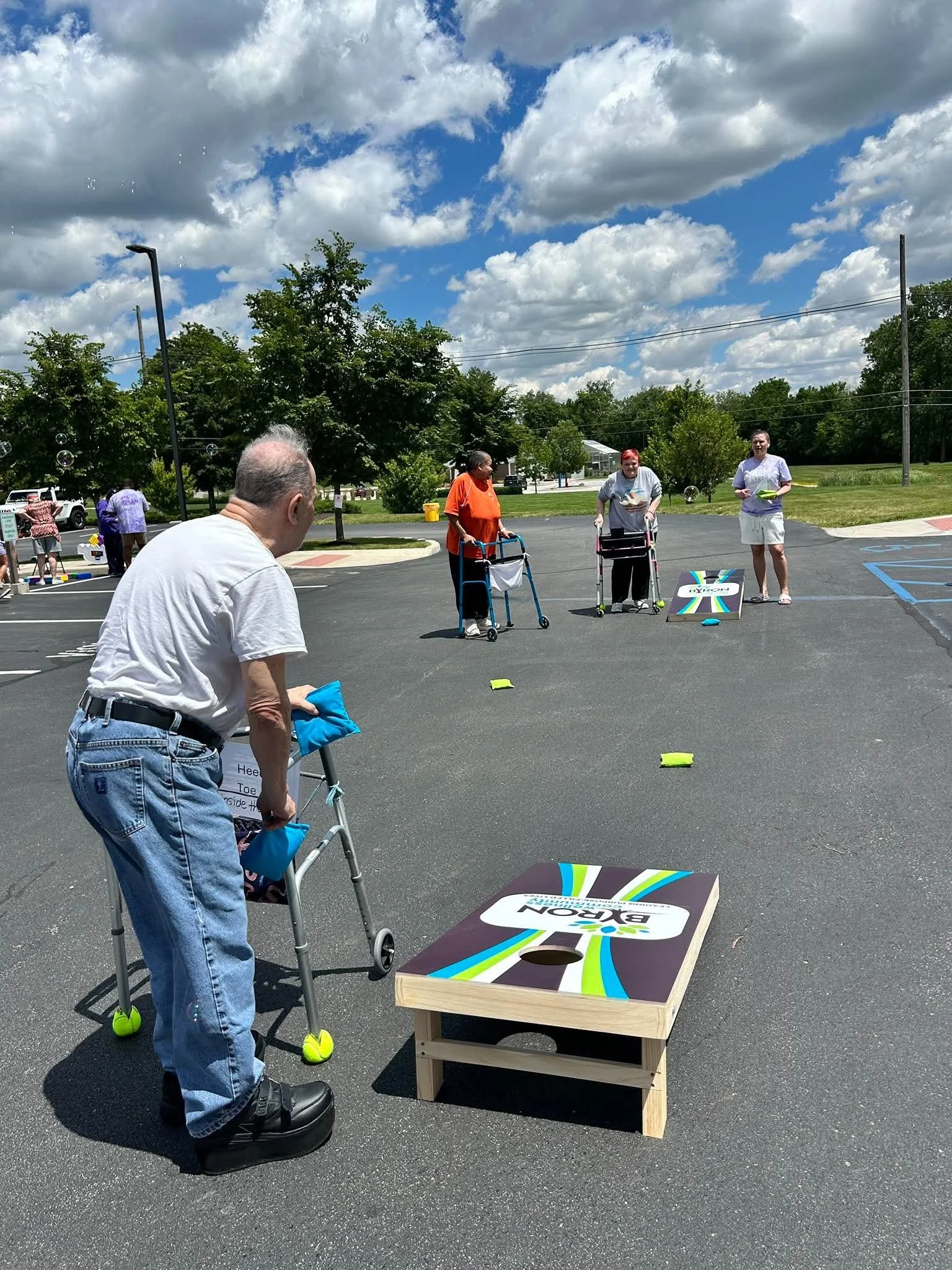 People playing cornhole outdoors on a sunny day with blue skies and clouds, some standing and some using walkers.