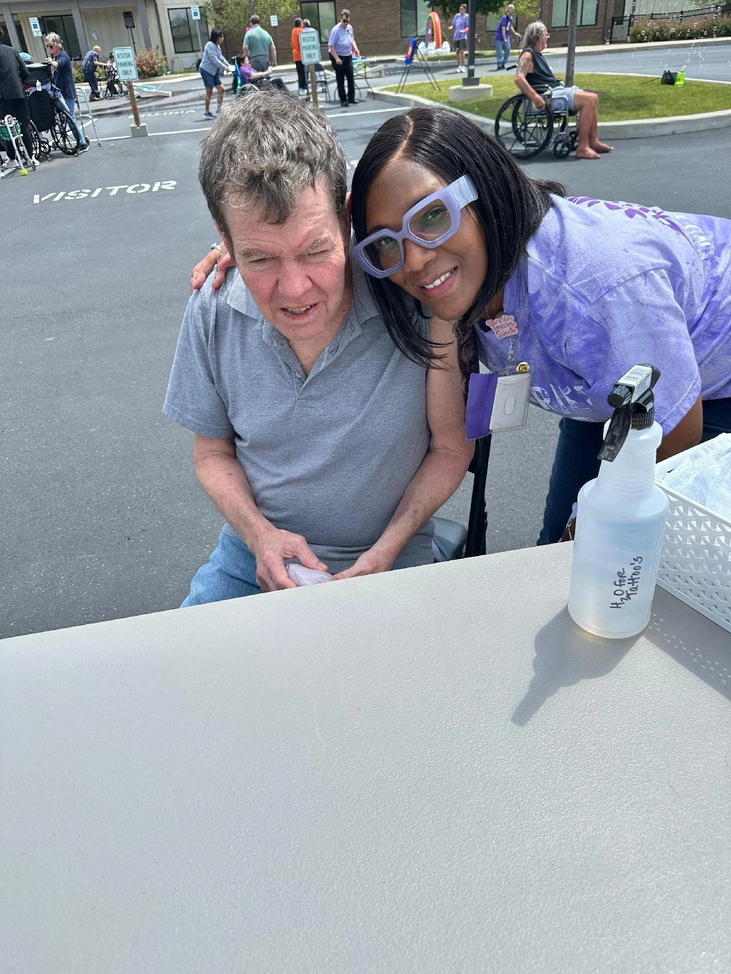 An older man with gray hair and a woman with black hair wearing large purple glasses sit together at a table outdoors. The woman is smiling and leaning toward the man, who has his eyes closed. A bottle of hand sanitizer labeled 'H2O for Matios' is on the table in front of them. In the background, there are several people, some in wheelchairs, gathered in a parking lot near a building with signs.