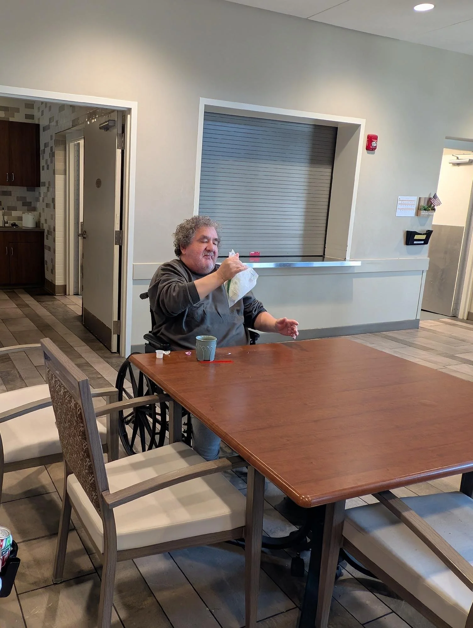 A man with curly gray hair and a gray beard sitting at a wooden table, holding a small bag of salad and a fork, with a ceramic mug in front of him. The setting appears to be a casual dining area in a cafeteria or restaurant.