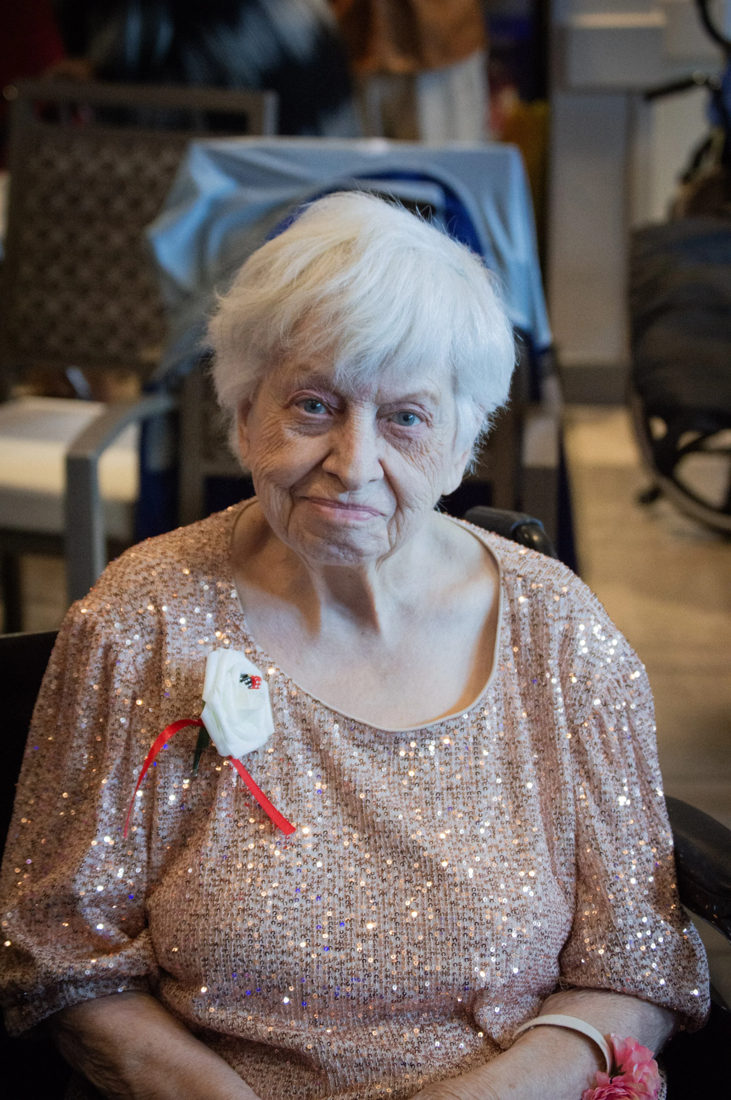 An elderly woman with white hair, wearing a sparkly peach-colored dress, sitting indoors at a celebration or event. She has a white flower corsage with a red ribbon on her dress and a pink flower wrist corsage. The background shows a blurred room wit