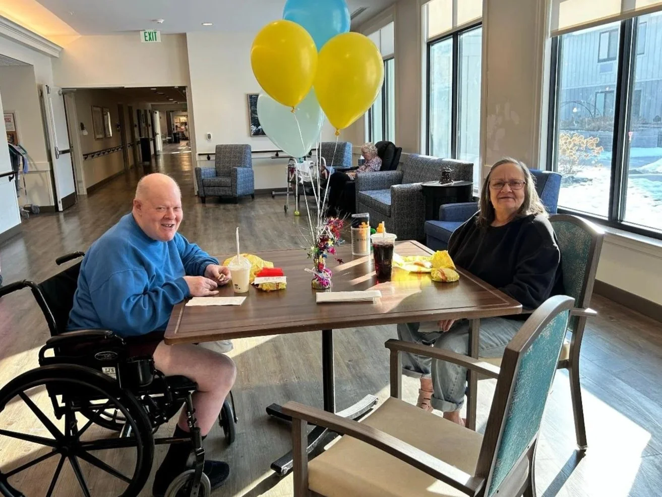 An elderly man in a wheelchair and an elderly woman sitting at a table with balloons, food, and drinks in a well-lit common area of a nursing home or assisted living facility.