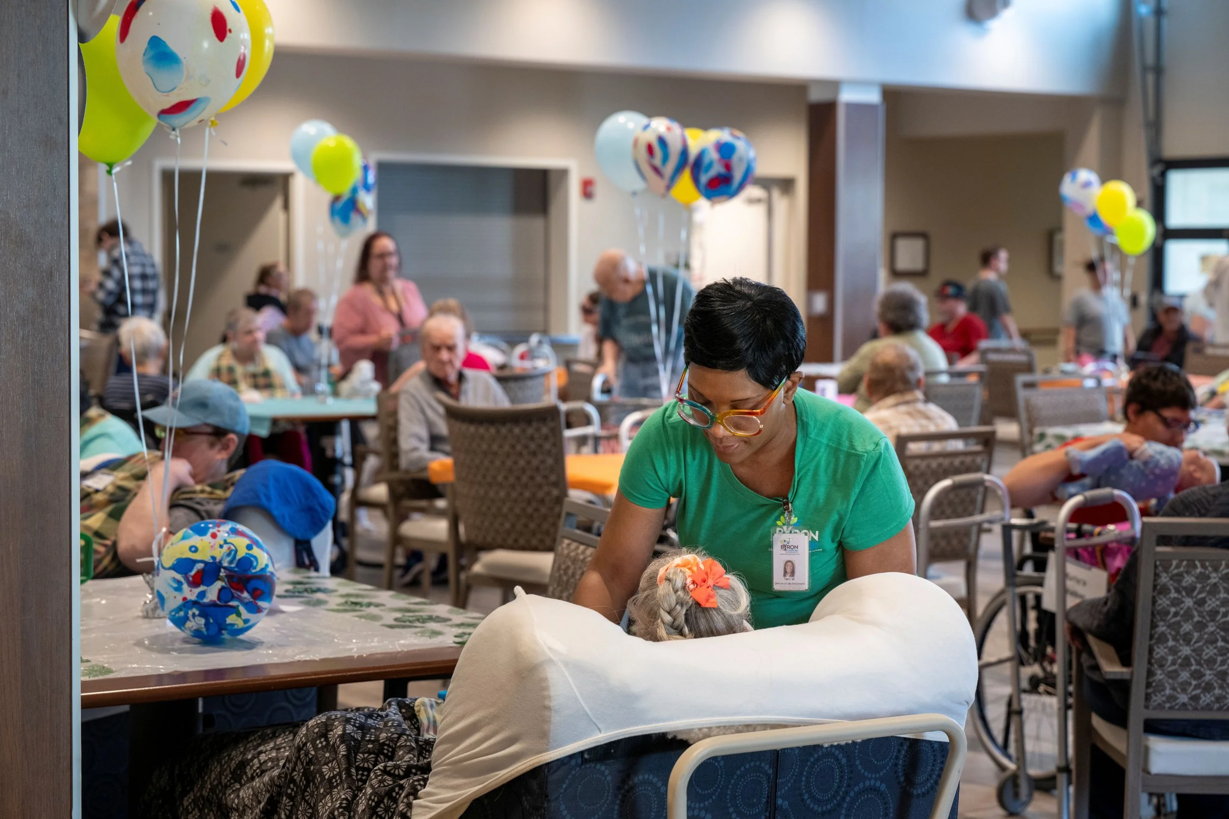 A group of elderly people sitting at tables in a large room decorated with colorful balloons, with a woman in a green shirt assisting a child.