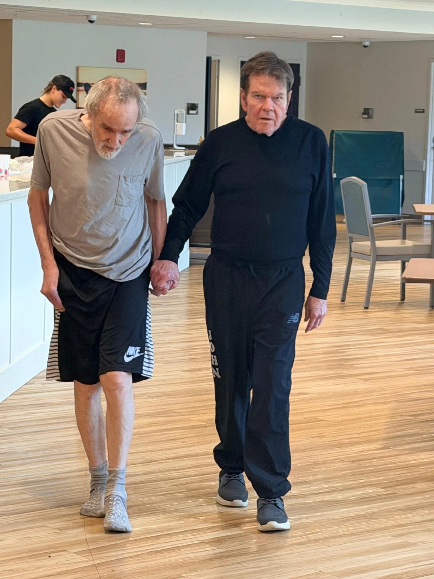 Two elderly men, one with grey hair and a beard, and the other with dark hair, are walking hand in hand in an indoor space. The man with grey hair is looking down, while the other man is looking directly at the camera with a serious expression. A you