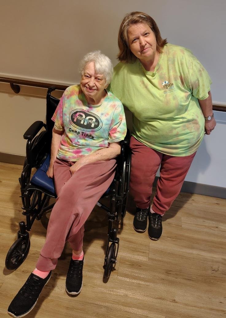 A smiling elderly woman in a wheelchair and a middle-aged woman standing next to her, both wearing colorful tie-dye shirts, in an indoor setting with wooden flooring and a beige wall.