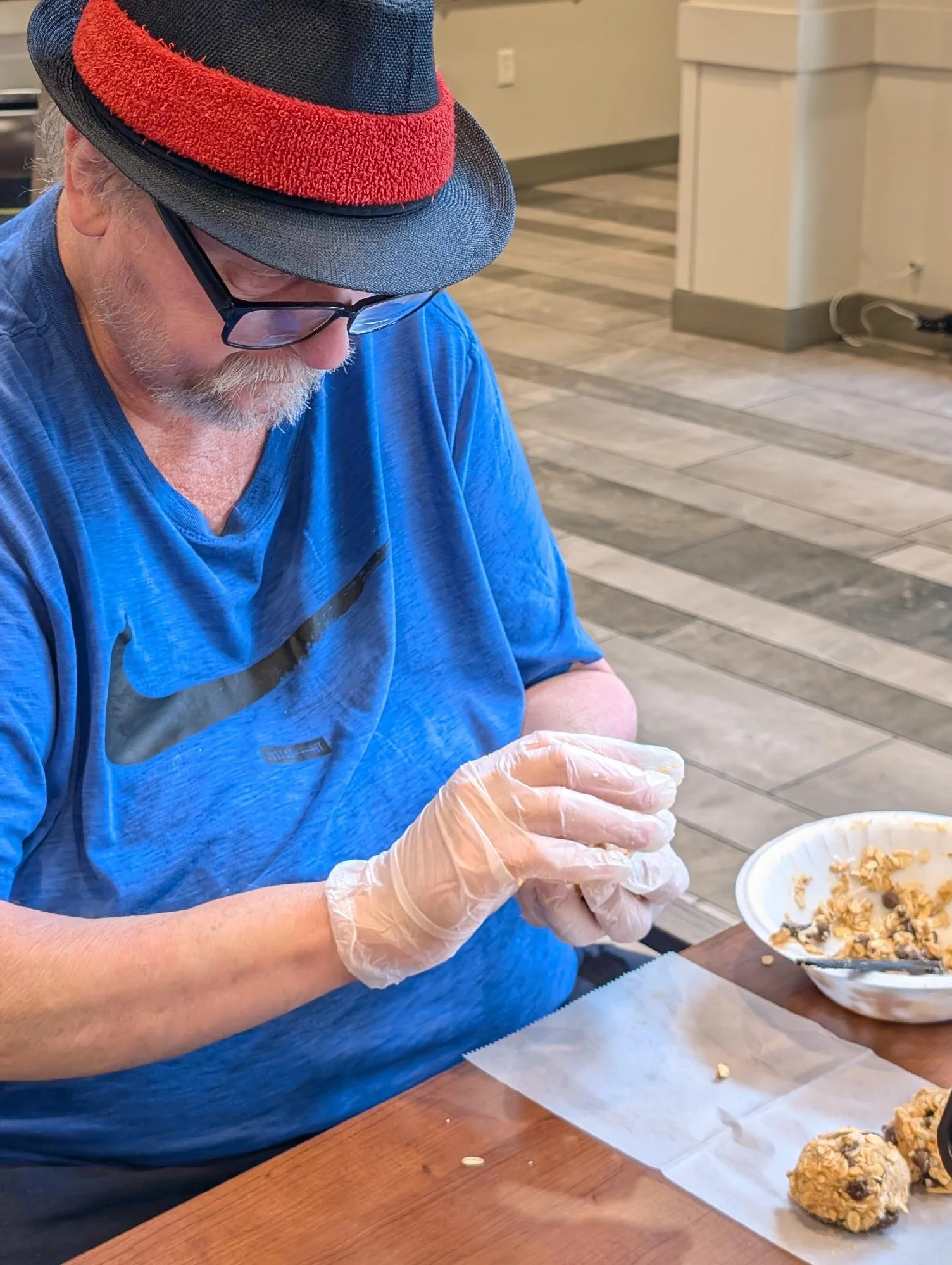 A man wearing glasses, a blue shirt, a black hat with a red band, and a plastic glove on his right hand is preparing snacks at a restaurant or dining area. He is holding a piece of food over a bowl of cereal or granola. There are more snack balls on 