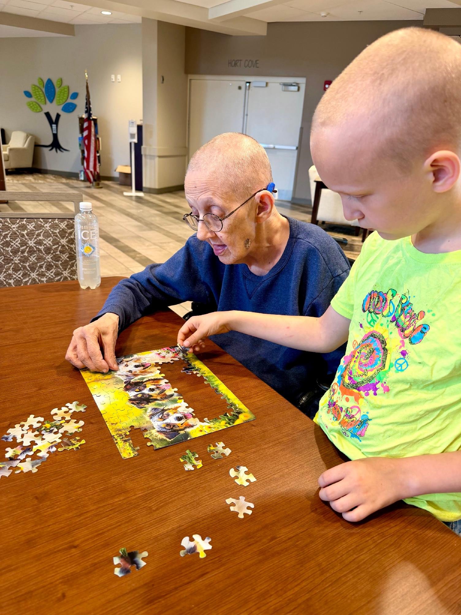 An elderly woman with glasses and a young boy with a buzz cut working on a puzzle with dog images at a wooden table.