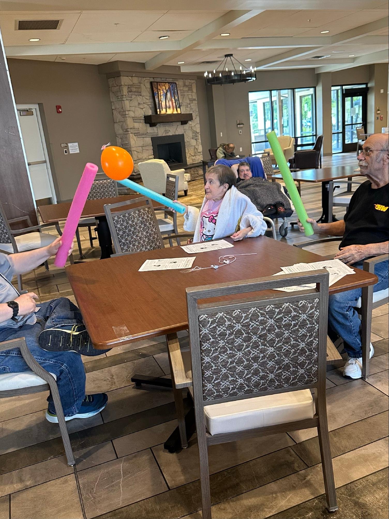 People sitting at a table in a lounge, playing with foam sticks and a balloon, with a fireplace and large windows in the background.