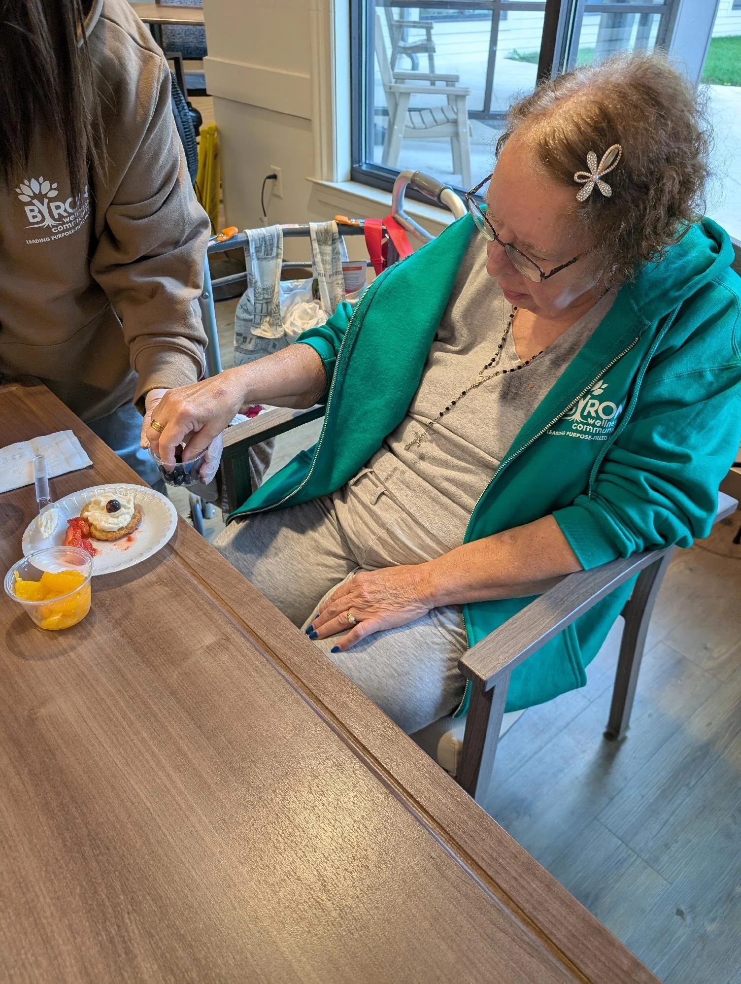 An elderly woman with glasses, curly hair, and a butterfly hair clip, wearing a green jacket and sitting in a wheelchair, is reaching for food on a plate on the table in front of her. A caregiver stands nearby.