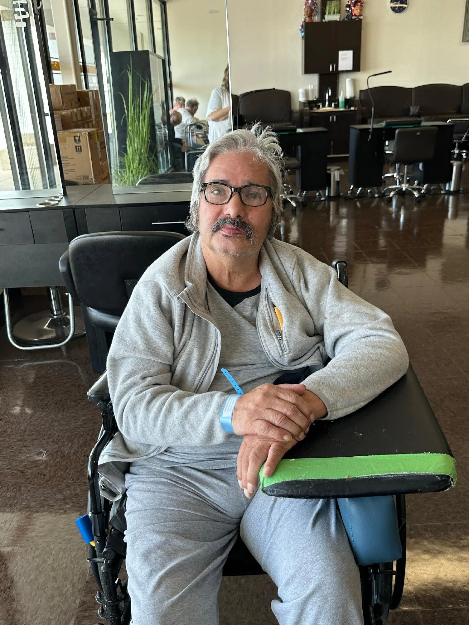 A man with gray hair, a mustache, and glasses sitting in a wheelchair inside a building, with a black table in front of him and wearing a light gray jacket and pants.