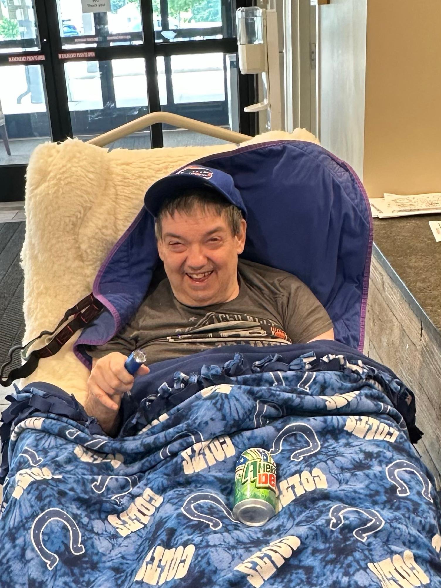 A man smiling while lying in a hospital bed, holding a blue object, with a Mountain Dew soda can resting on his bedspread. He wears a blue cap and a gray T-shirt. The bed has a pirate-themed blanket with blue and white designs. Background shows hospi