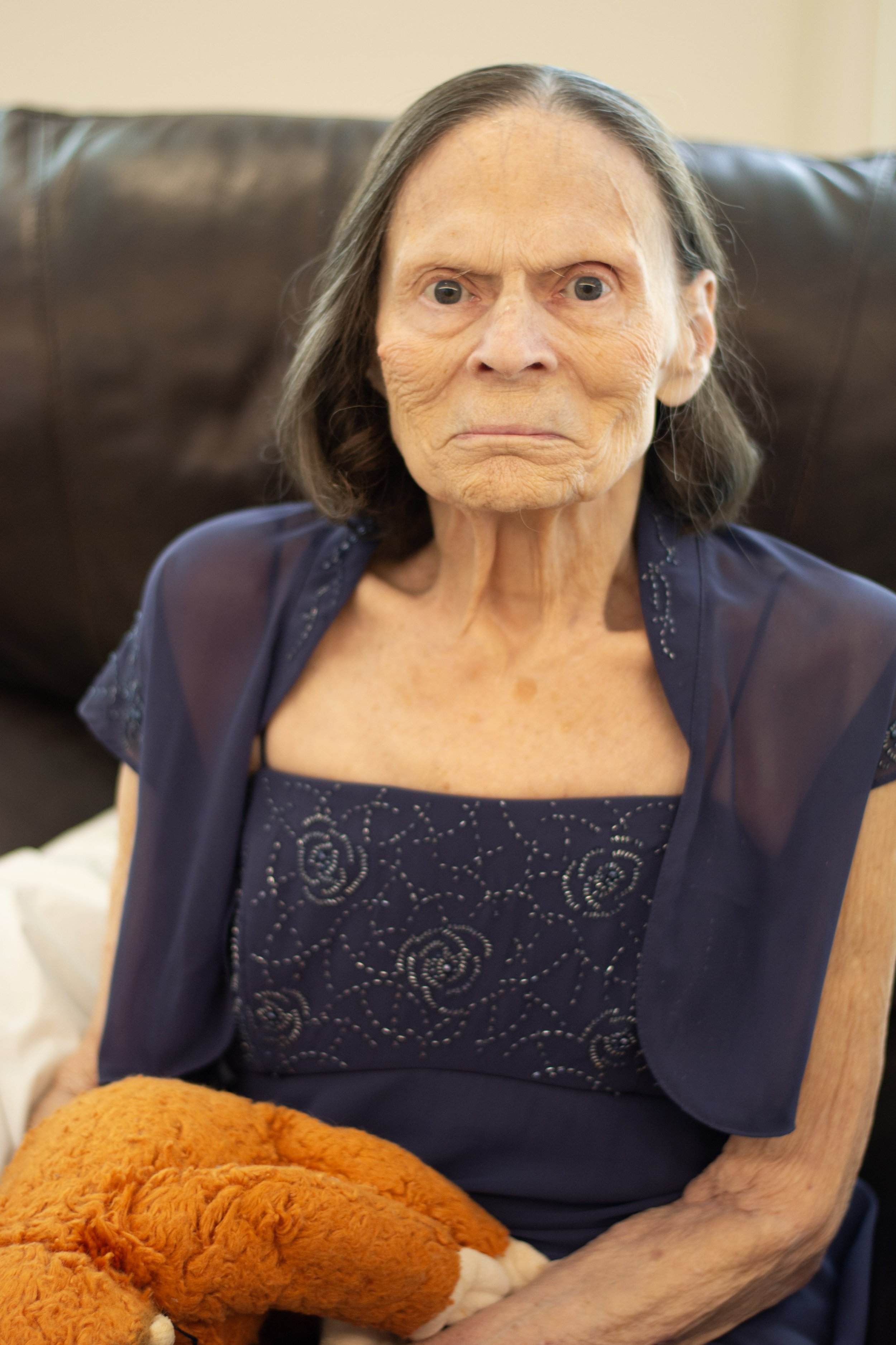 An elderly woman with gray hair sitting on a dark brown leather chair, wearing a navy blue dress with floral embroidery, holding a brown plush teddy bear.