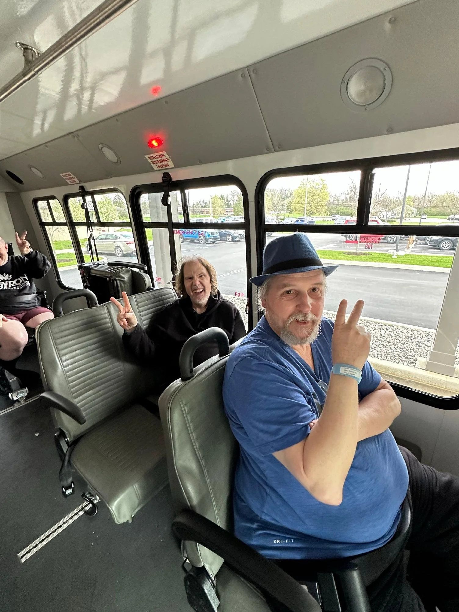 Three men sitting on the bus, two making peace signs and smiling at the camera. The bus has large windows and gray seats, with an outside view of parked cars and trees.