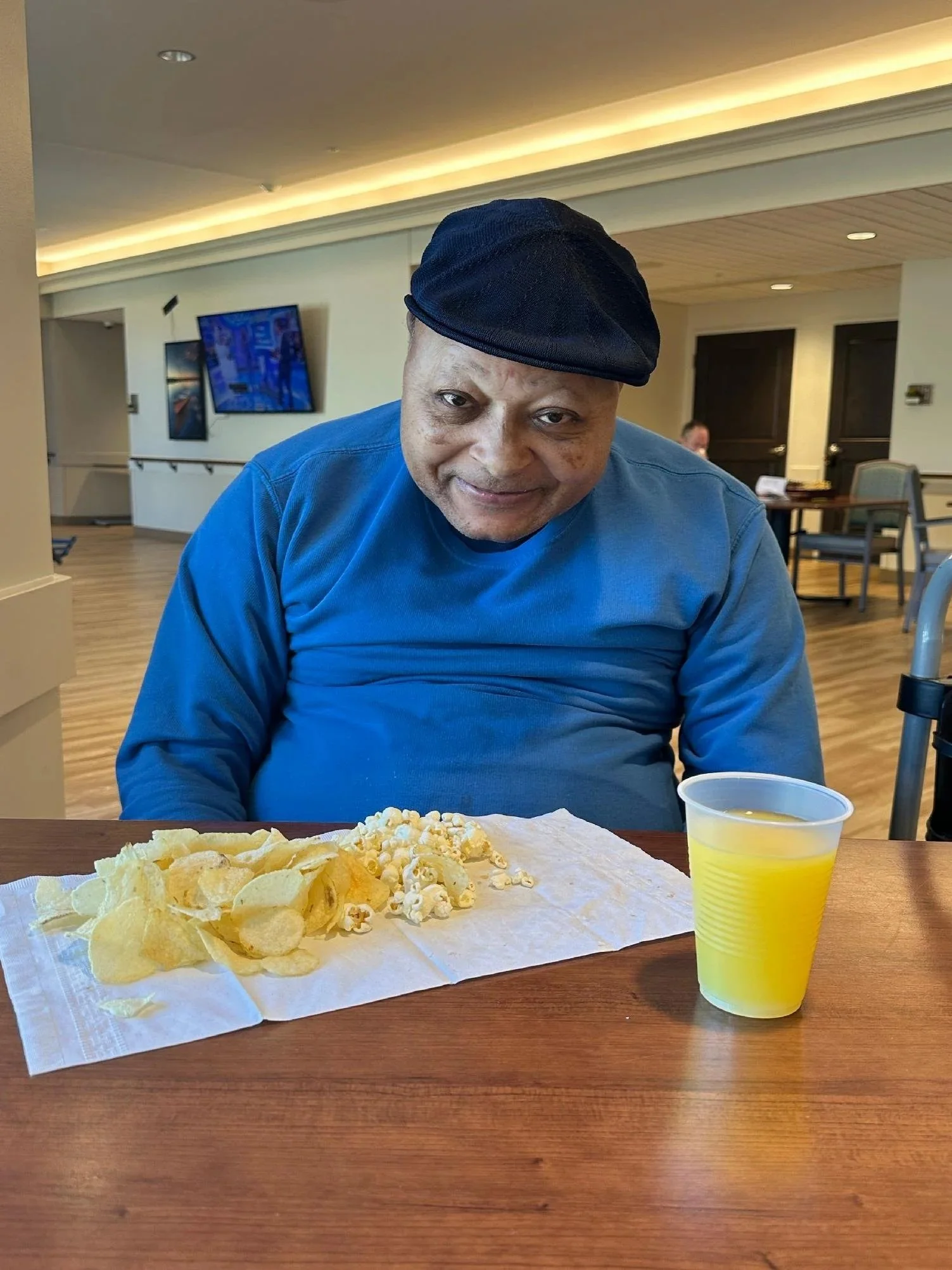 A man wearing a black hat and blue shirt sitting at a table with potato chips, popcorn, and a yellow drink in a clear plastic cup, in a well-lit indoor setting.