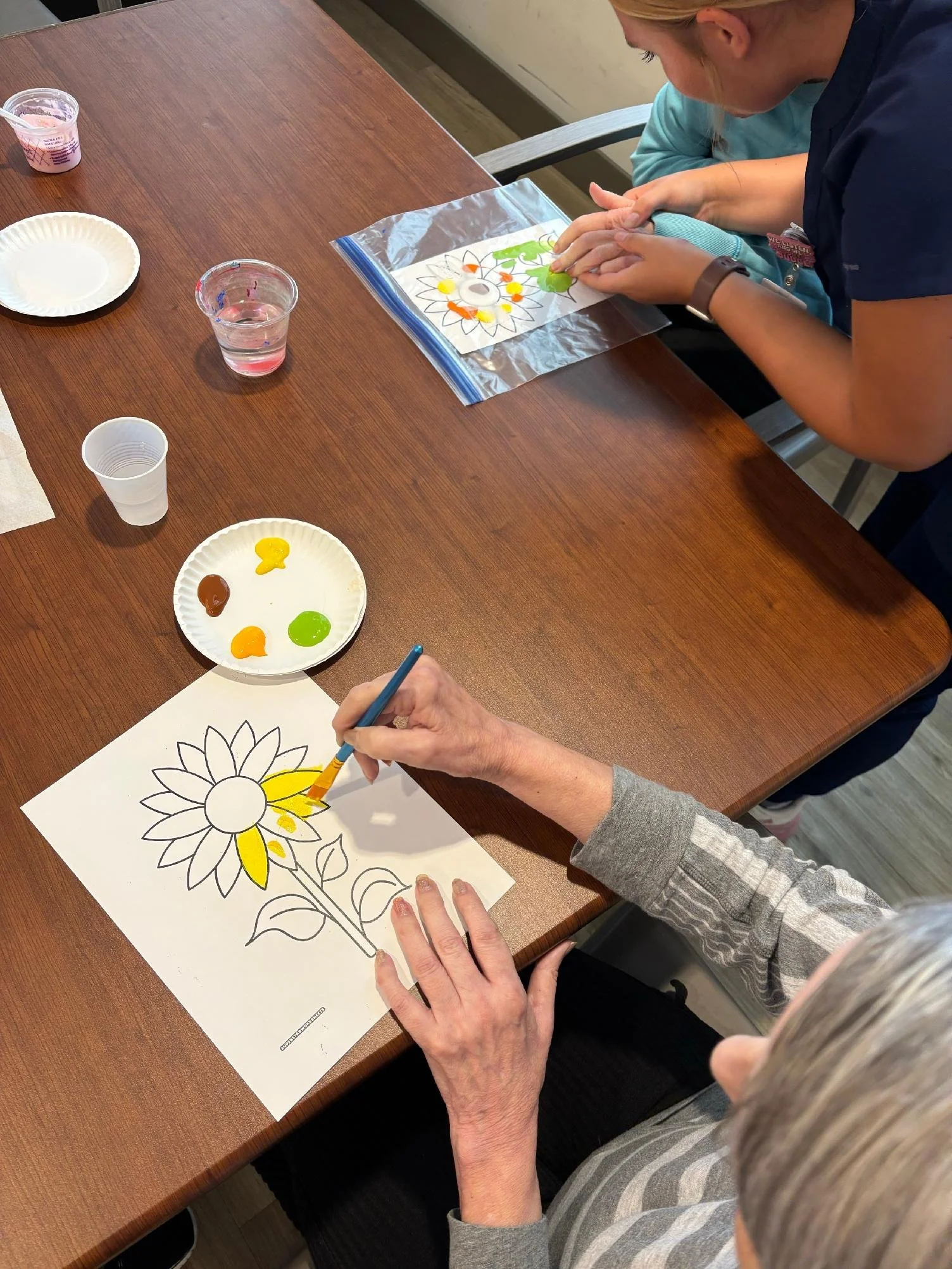 Older woman coloring a sunflower on a printed sheet with paint, surrounded by paint palette, glass of water, and paper plates on a wooden table.