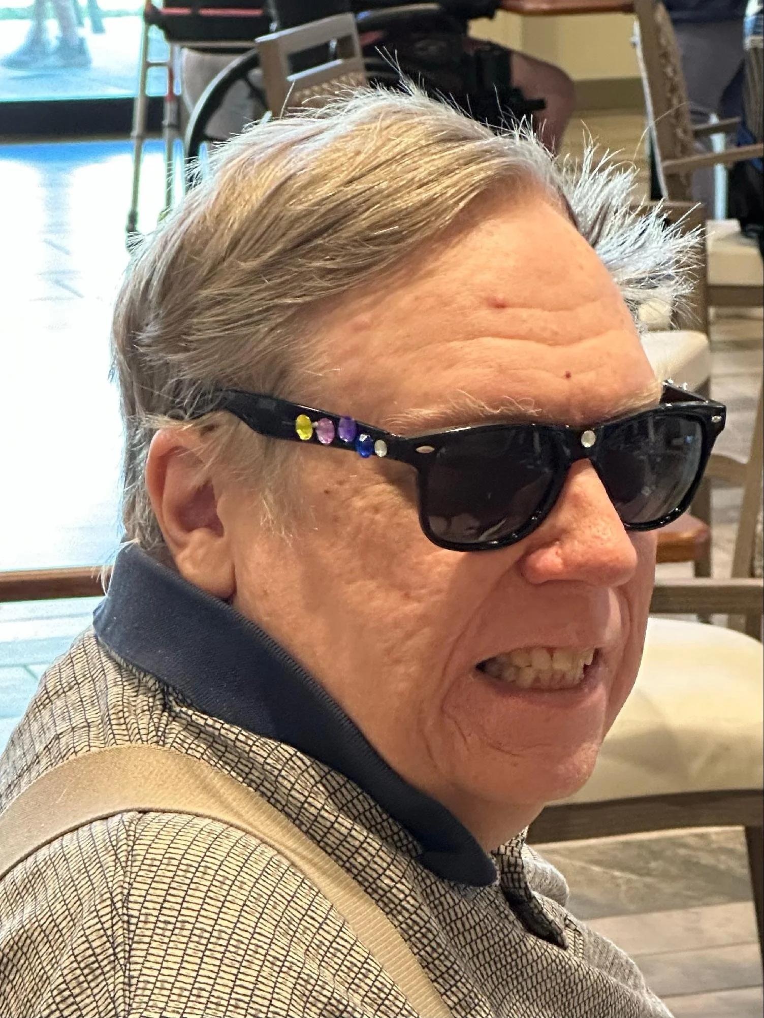 Close-up of an elderly man smiling, wearing sunglasses with decorative stones, a beige jacket with a checkered pattern, and a black shirt underneath, inside a cafe or restaurant.