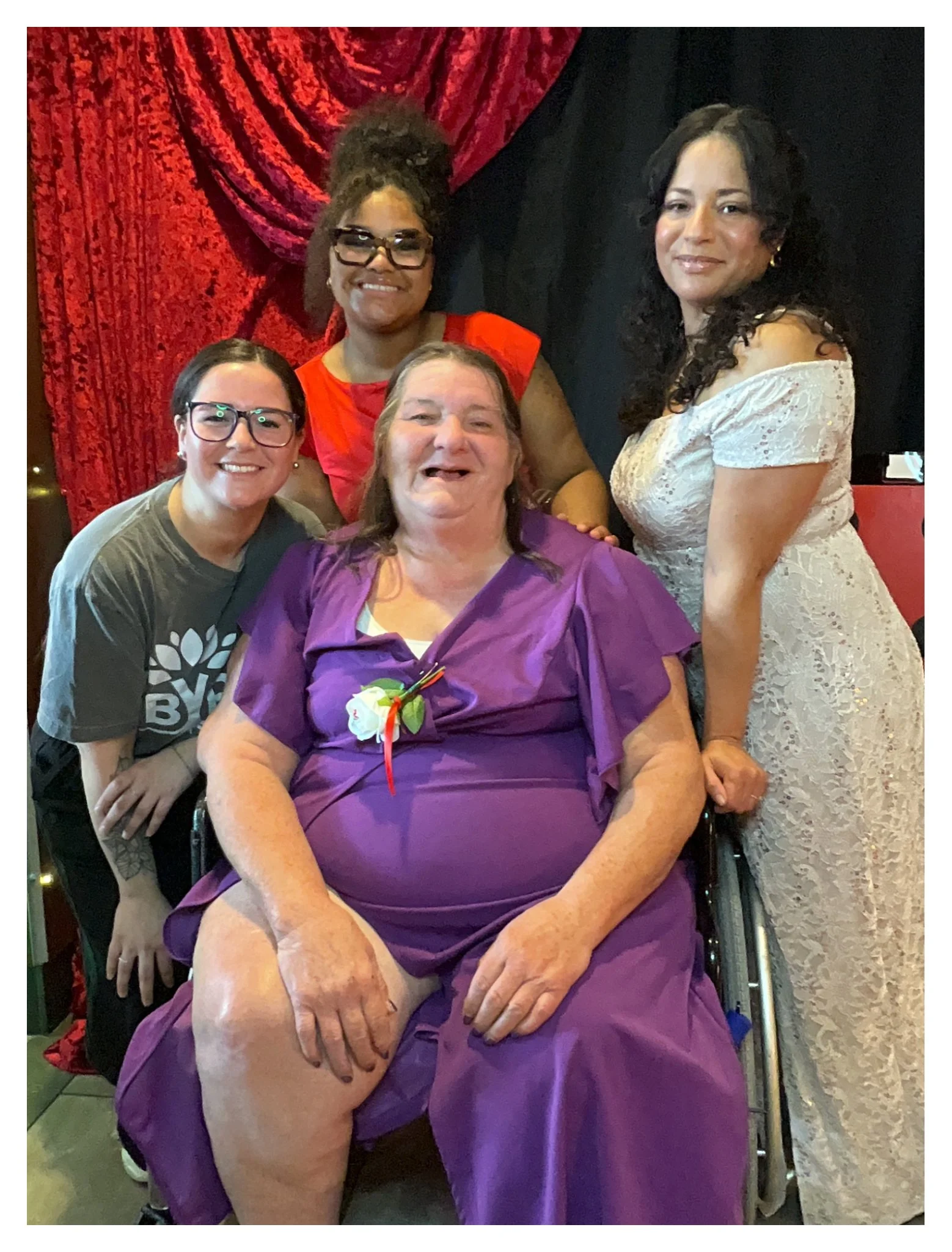 Group of five women posing together at a celebration. One woman in the front is seated in a wheelchair, wearing a purple dress with a corsage. Four women stand behind her, smiling, with a red curtain backdrop and black background behind them.