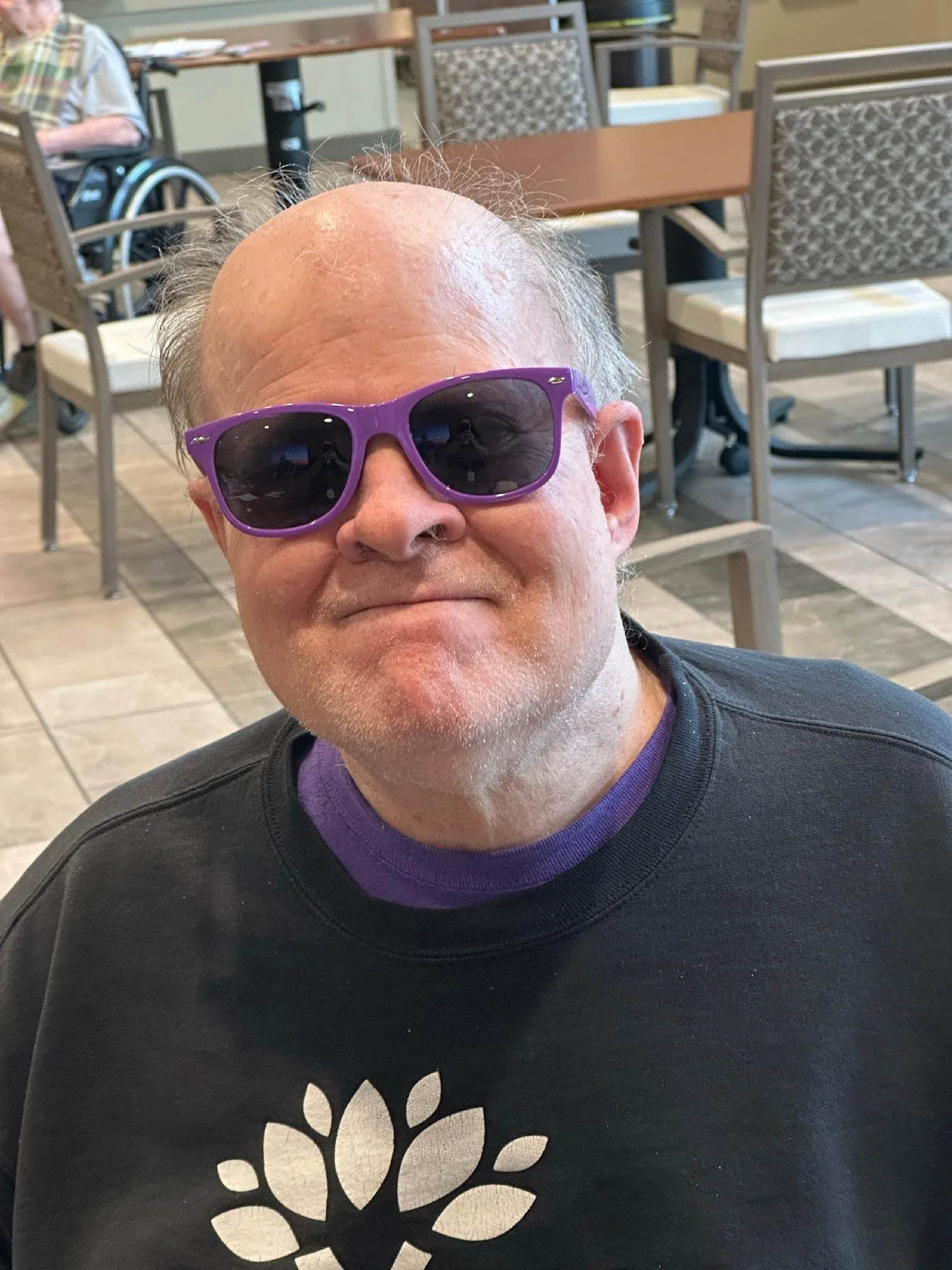 A smiling man wearing purple sunglasses and a black shirt with a white floral pattern in a restaurant or cafe.