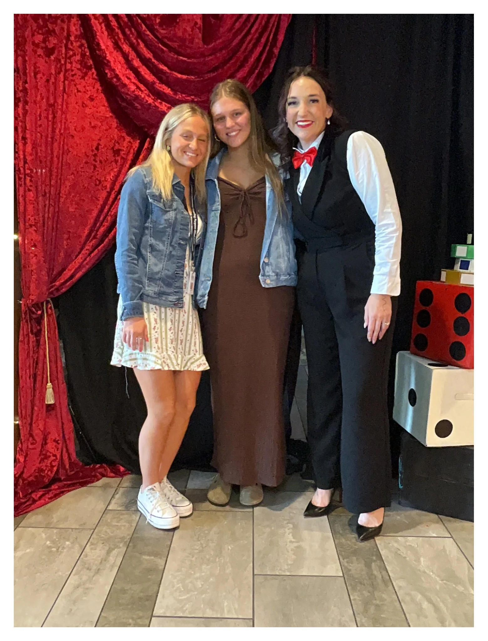 Three women posing together in front of a red velvet curtain with black background. The woman on the left has long blonde hair, wearing a denim jacket, a white dress with a striped pattern, and white sneakers. The woman in the middle has long light b