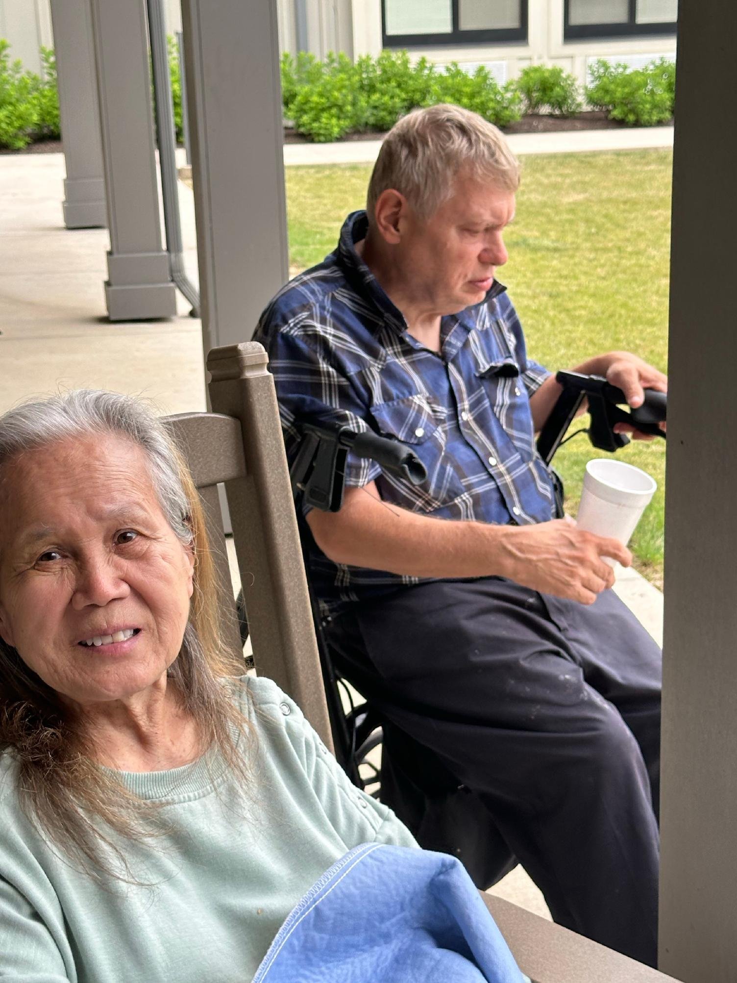 Smiling elderly woman with long gray hair in the foreground, woman in a wheelchair with short gray hair looking at phone in the background, sitting outside on patio with grass and bushes behind.