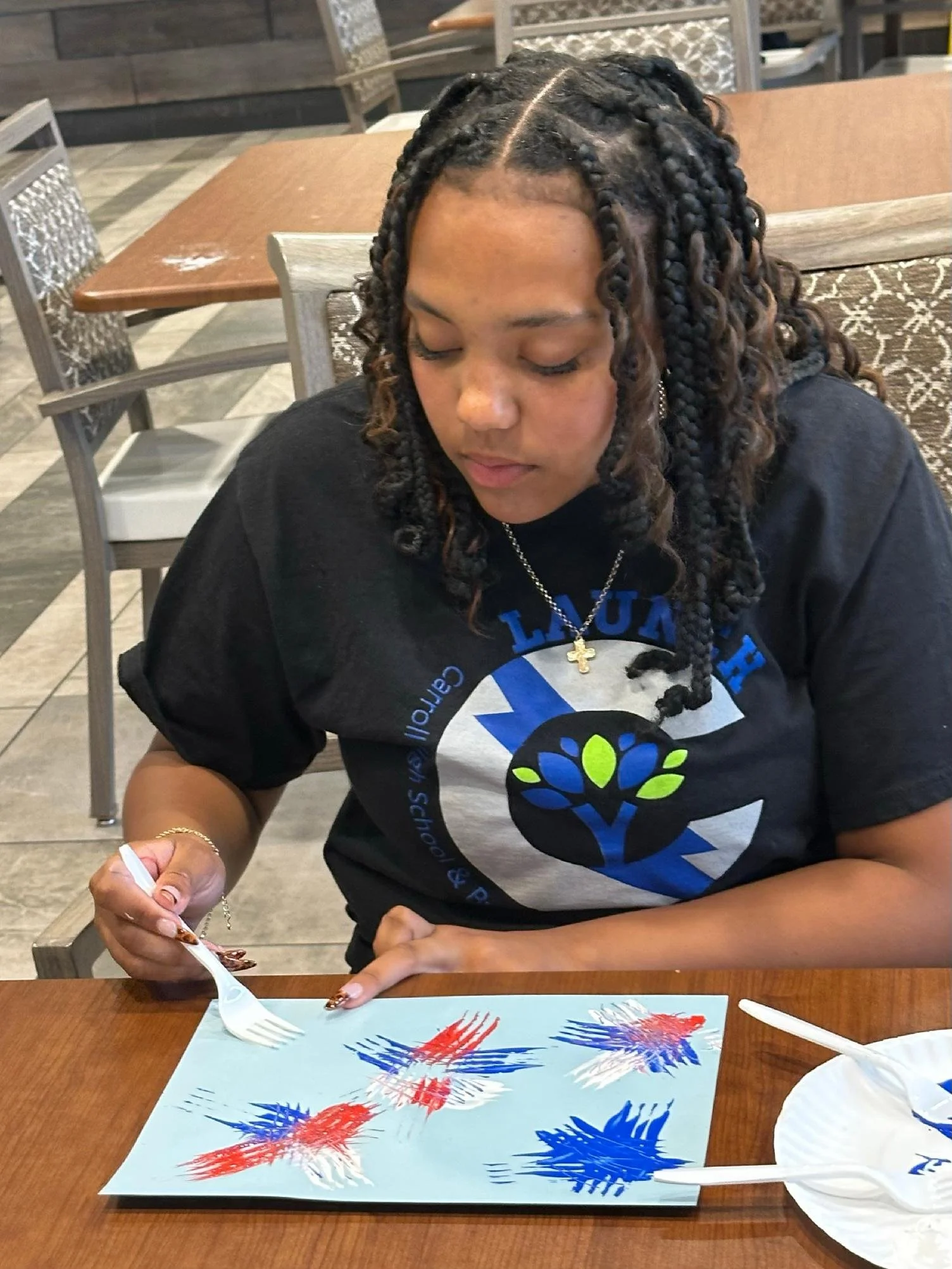 A young woman with braided hair is sitting at a table, painting on a blue sheet of paper with red and blue paint. She is holding a fork with some paint on it, and there is a white disposable mask on the table.