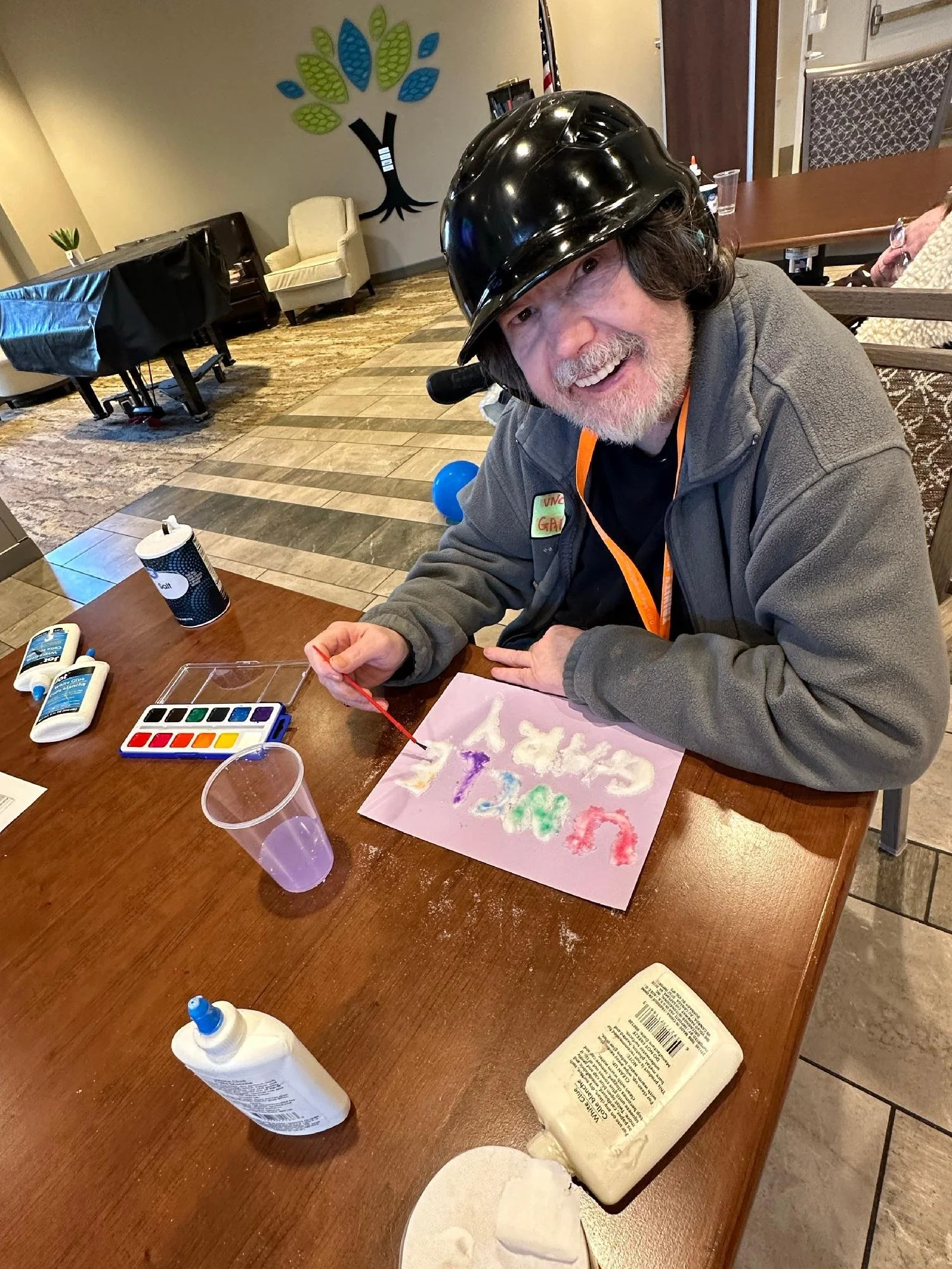 A man with a black helmet smiling at a table with watercolors, paint, and art supplies. The table has a pink paper with colorful watercolor smudges. In the background, there is a wall with a tree decoration, a beige armchair, and a table with a black