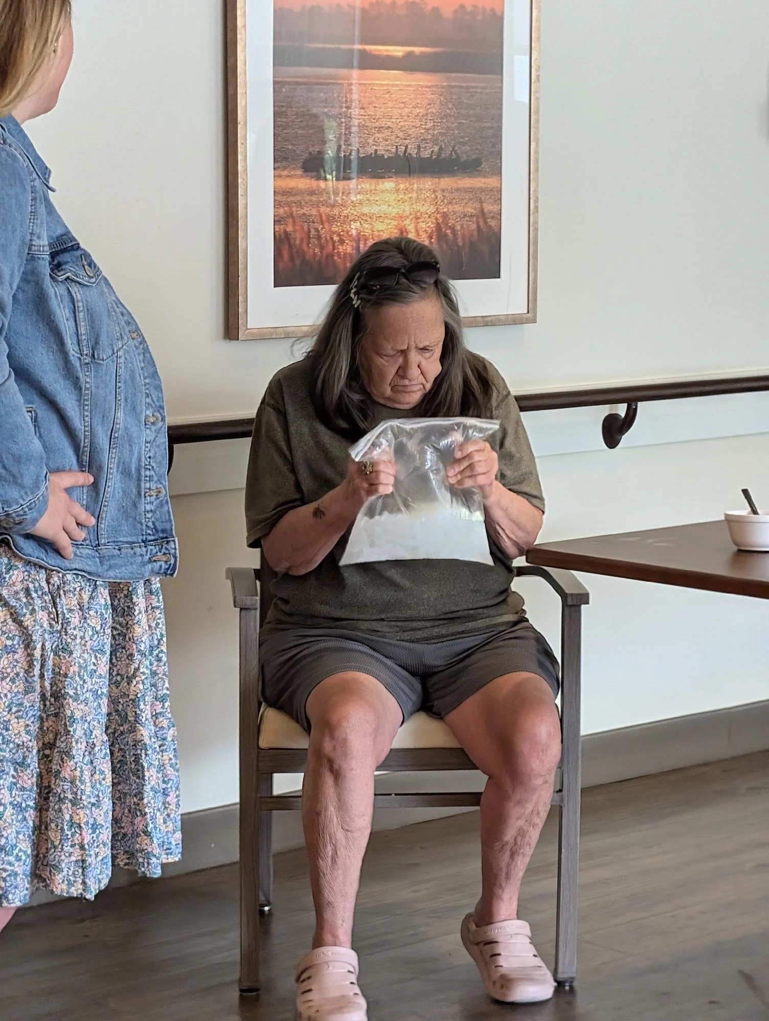 An elderly woman sitting in a chair, looking into a plastic bag, with a woman standing beside her. The scene is indoors, with a framed sunset over a body of water on the wall behind them. The seated woman is wearing shorts and Crocs.