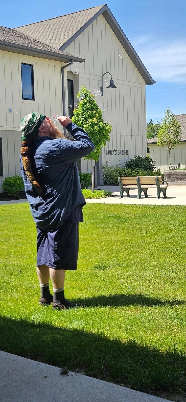 A man with a beard, wearing a knit hat, blue long-sleeve shirt, dark shorts, and socks, standing on a grassy lawn, blowing bubbles in a sunny backyard with a white house labeled 'Laura's Garden', a tree, and park benches.