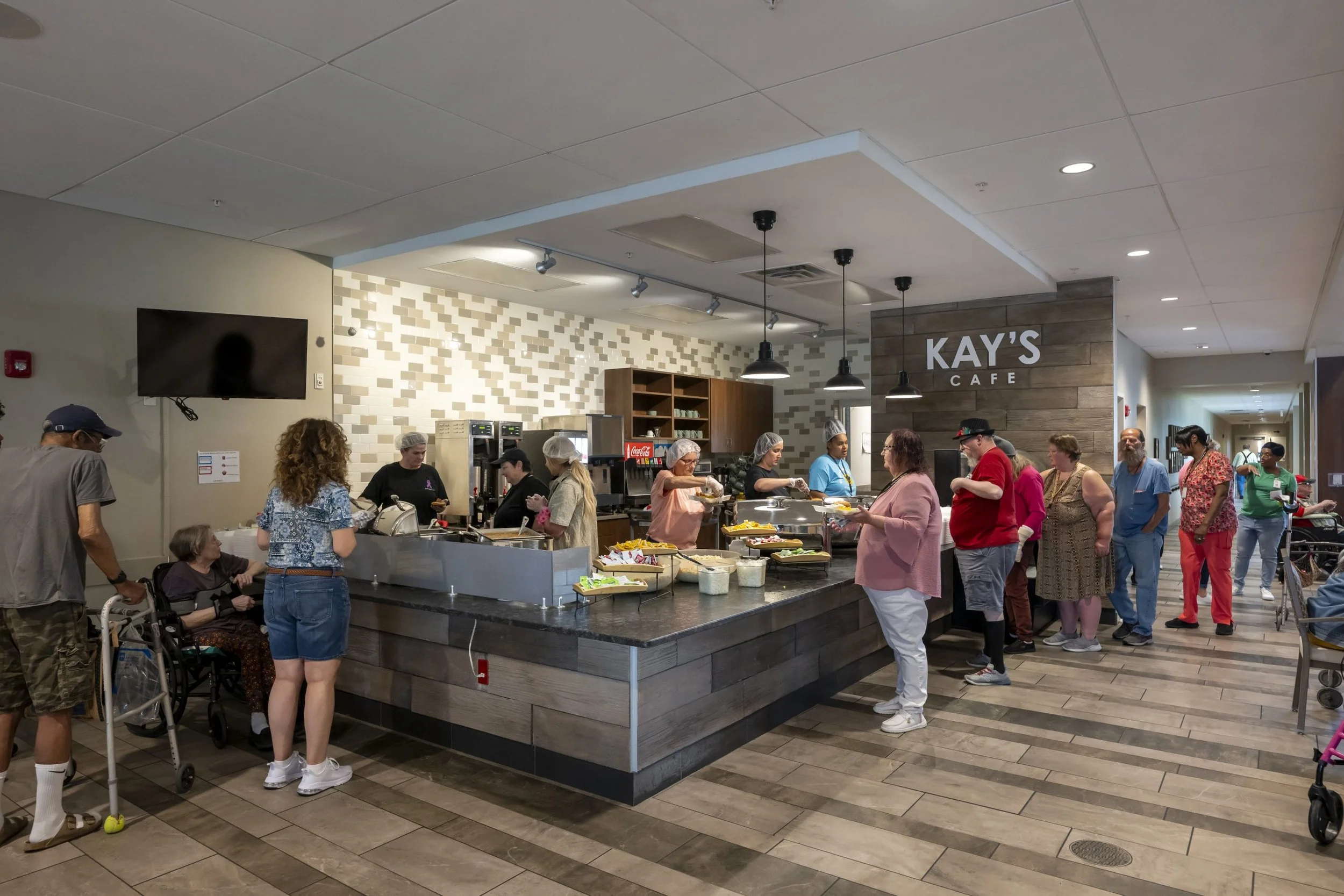 People lined up at a cafeteria serving food in a hospital or community center. There is a counter with food trays and servers, and several individuals are waiting to be served, including a woman with a walker and a man in a baseball cap.