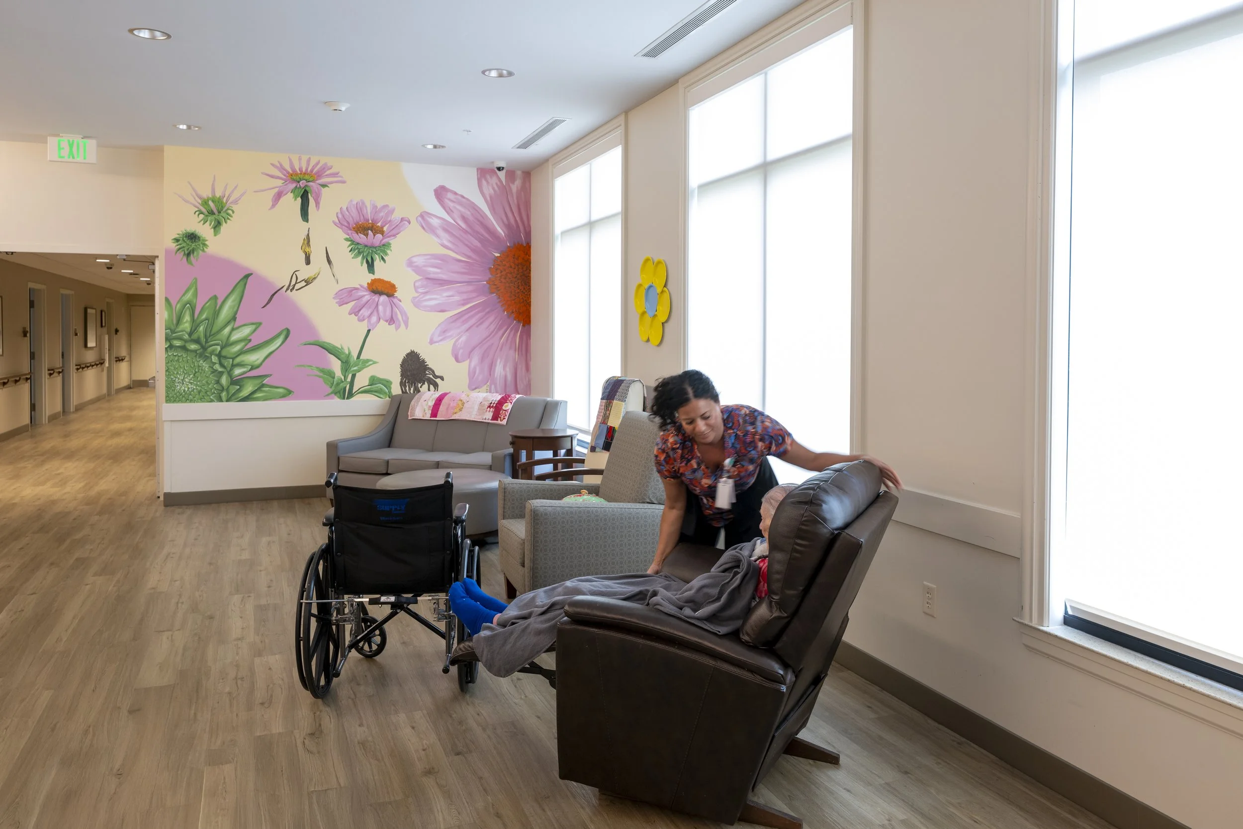 A woman in patterned scrubs helping a person in a wheelchair in a bright patient lounge with large windows and floral murals on the wall.