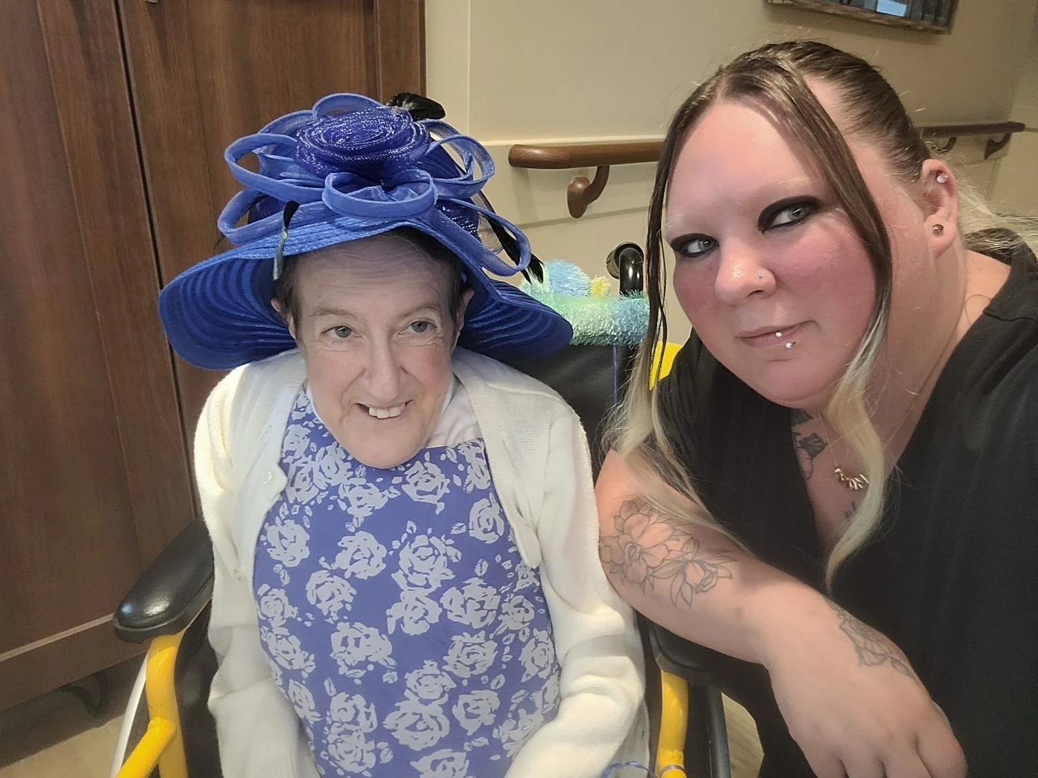 A woman and an elderly lady sitting together, both smiling and looking at the camera. The elderly lady is wearing a large, elaborate blue hat with ribbon details and a white cardigan. The woman has tattoos on her arm, shoulder-length blonde hair, and