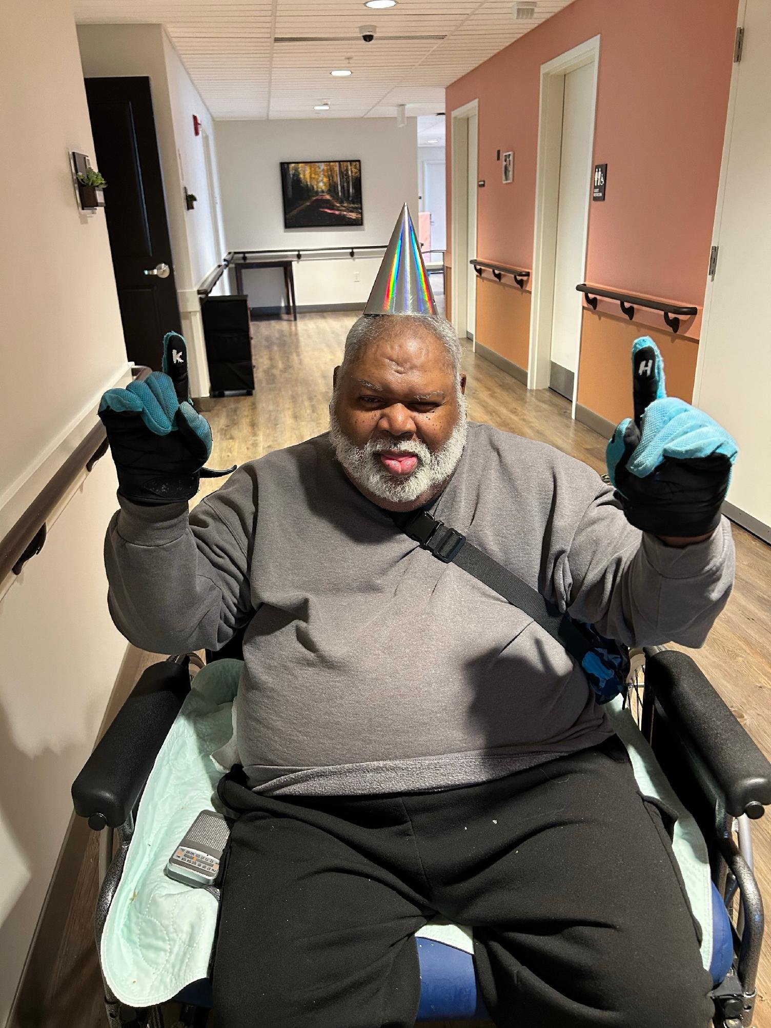 An elderly man with a gray beard wearing a party cone hat, gloves, a gray shirt, and black pants, sitting in a wheelchair in a hallway of a medical or care facility.