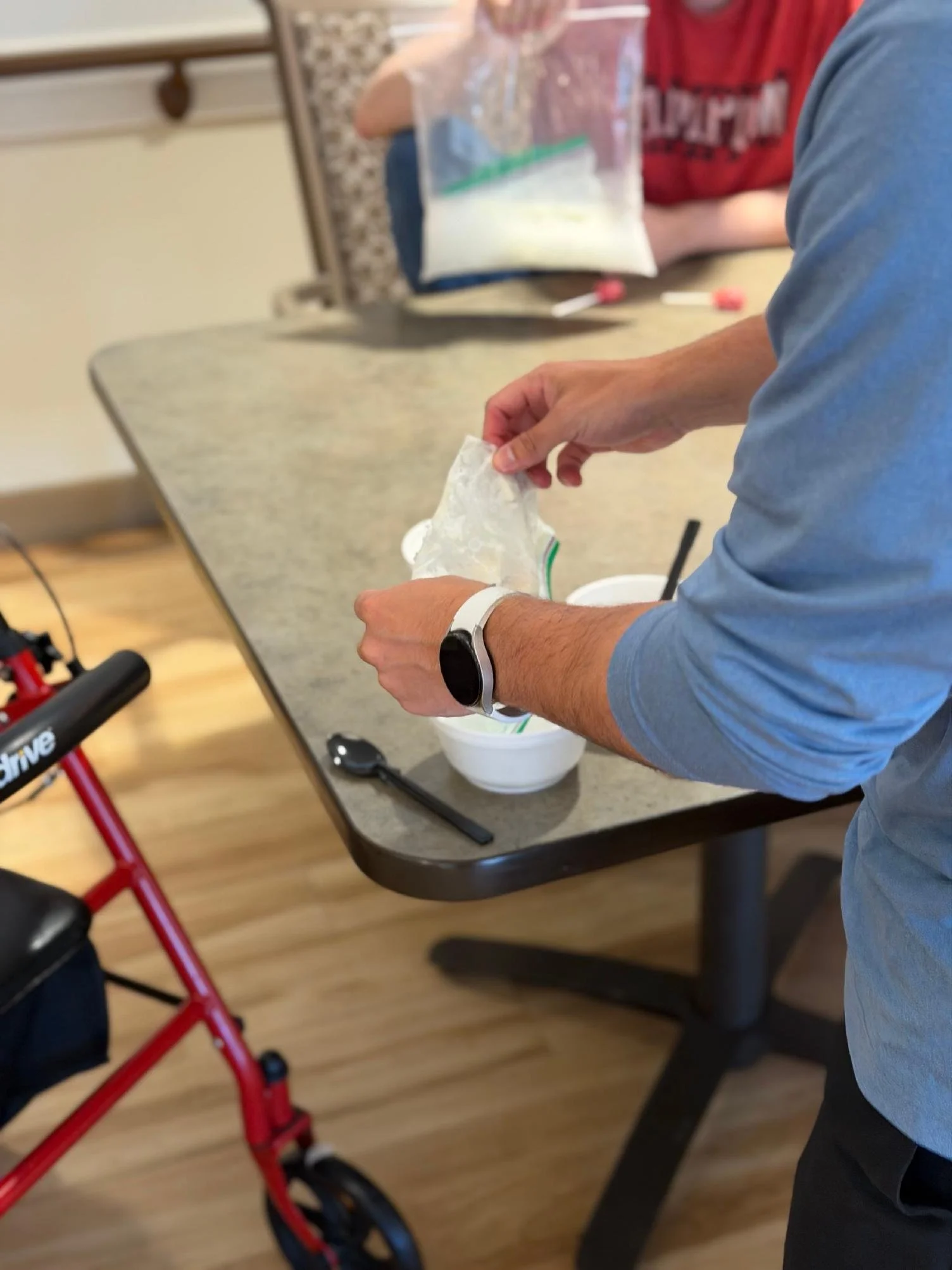 Person holding a small bag or packet over a container, with a walker nearby in a room with a table and a chair.