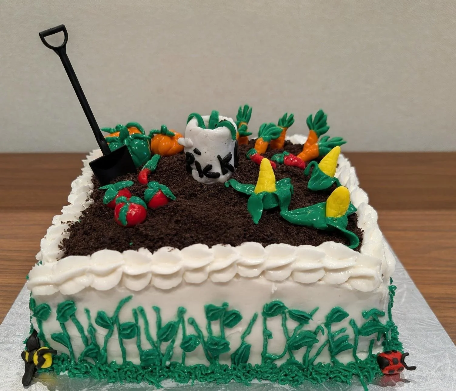 A square birthday cake decorated with a garden scene, including soil made of crumbled cookie crumbs, plastic carrots, a gardening shovel, and a white mug with black letters, topped with green icing plants and small plastic ladybug and bee decorations