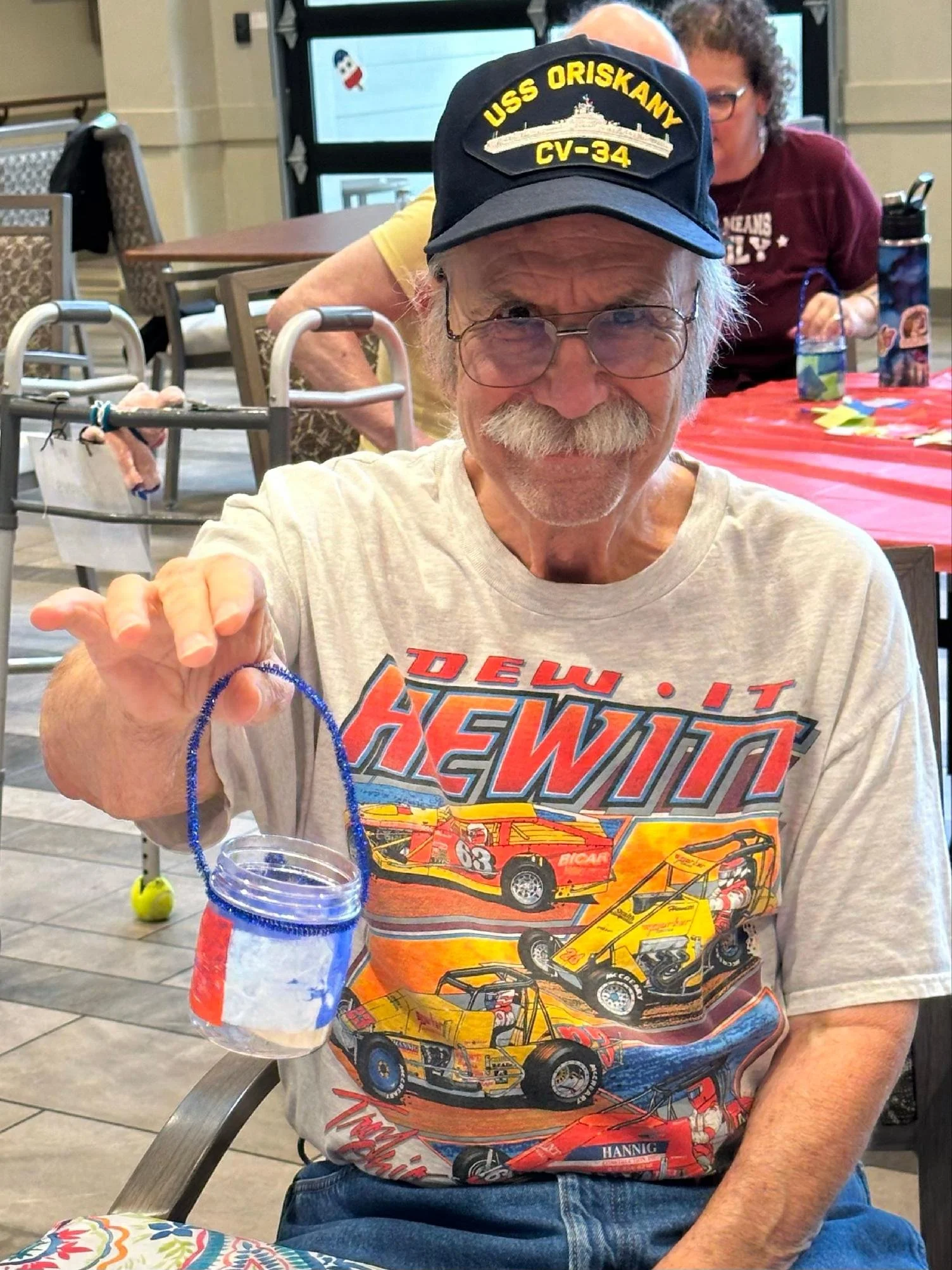 Older man with glasses and white hair, smiling, wearing a blue baseball cap with yellow embroidery, a graphics T-shirt with racing cars, and sitting at a table during a celebration or gathering.