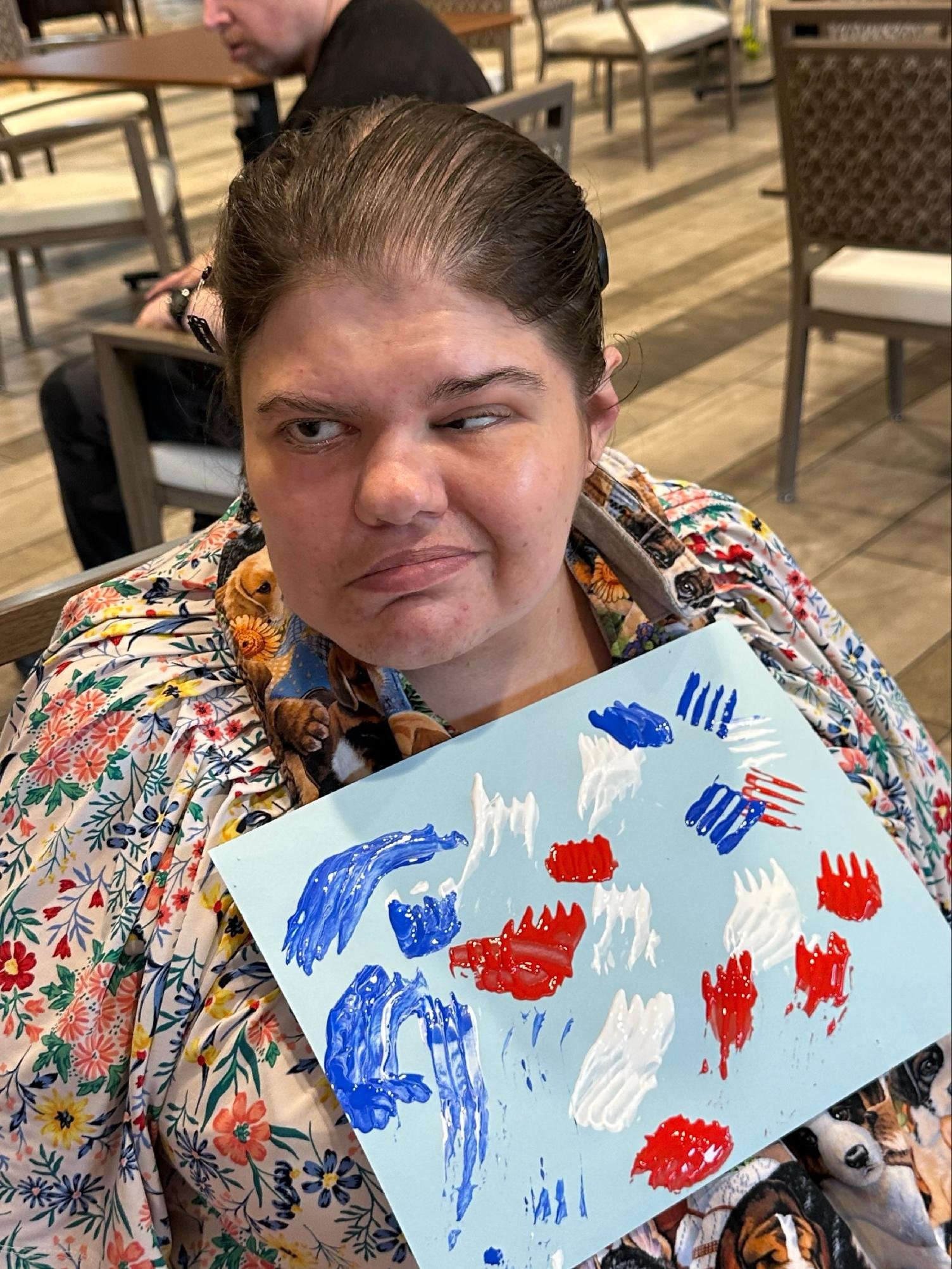 A woman with dark hair pulled back in a clip, wearing a floral shirt, is holding a canvas with blue, white, and red paint strokes. A man is sitting at a table in the background, looking down. The setting appears to be a restaurant or cafe with wooden