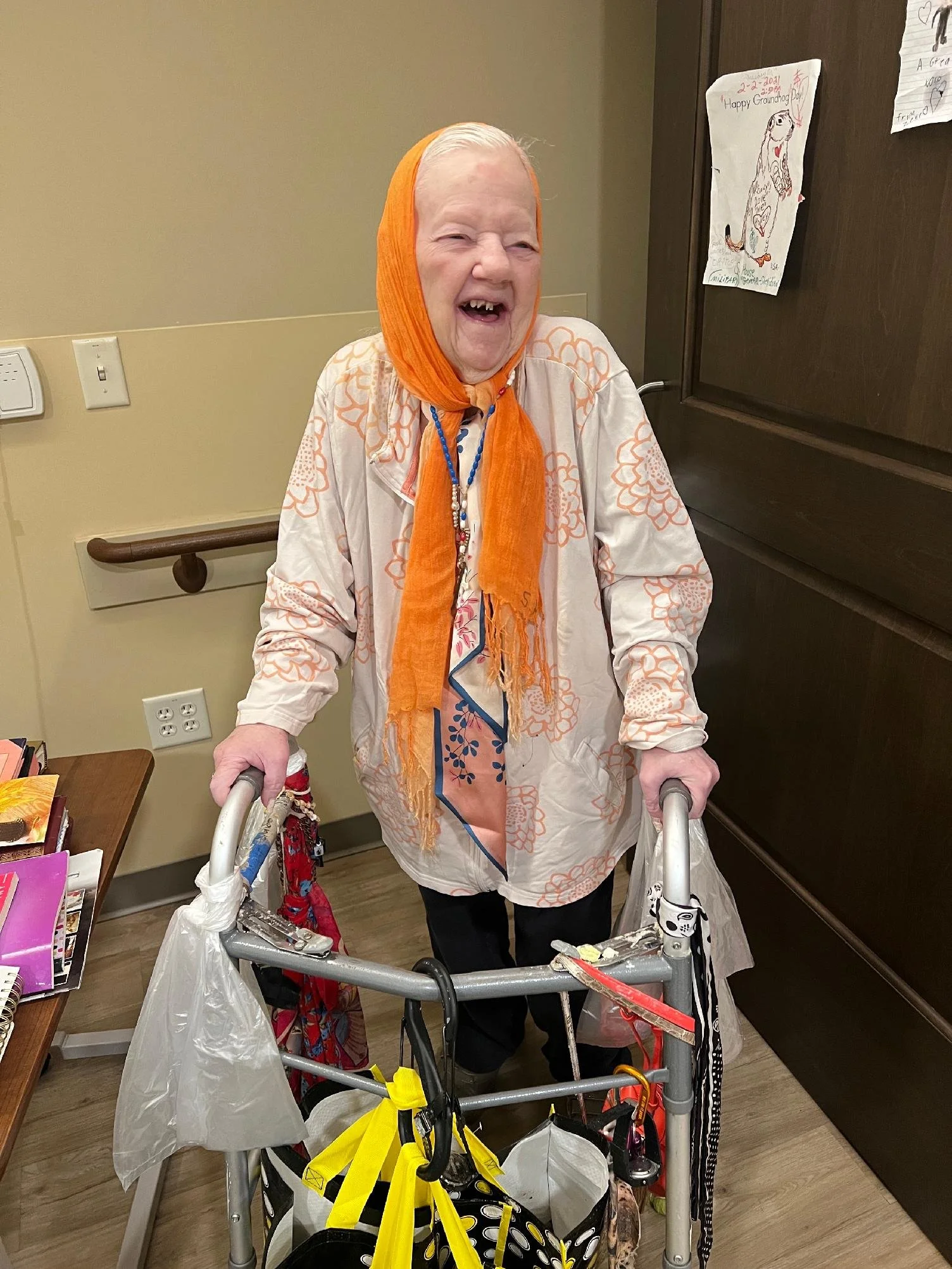 An elderly woman with a head covering, smiling and holding onto a walker, standing in a room with drawings on the wall.