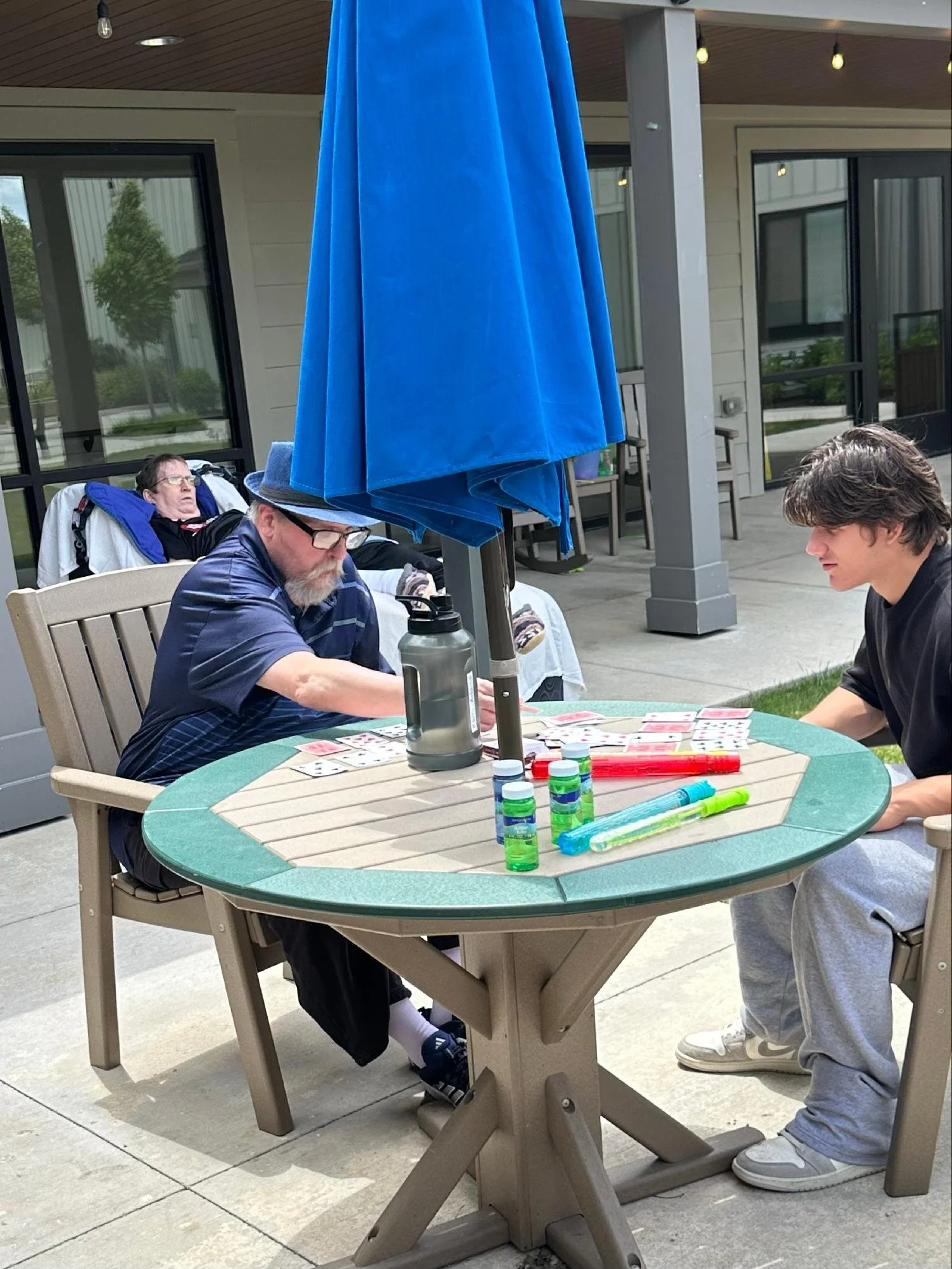 Three men are sitting and standing around a round outdoor table set for a game, with playing cards and water bottles. One man in a wheelchair is seated at the table, while two others are standing, interacting. There are large blue umbrellas providing