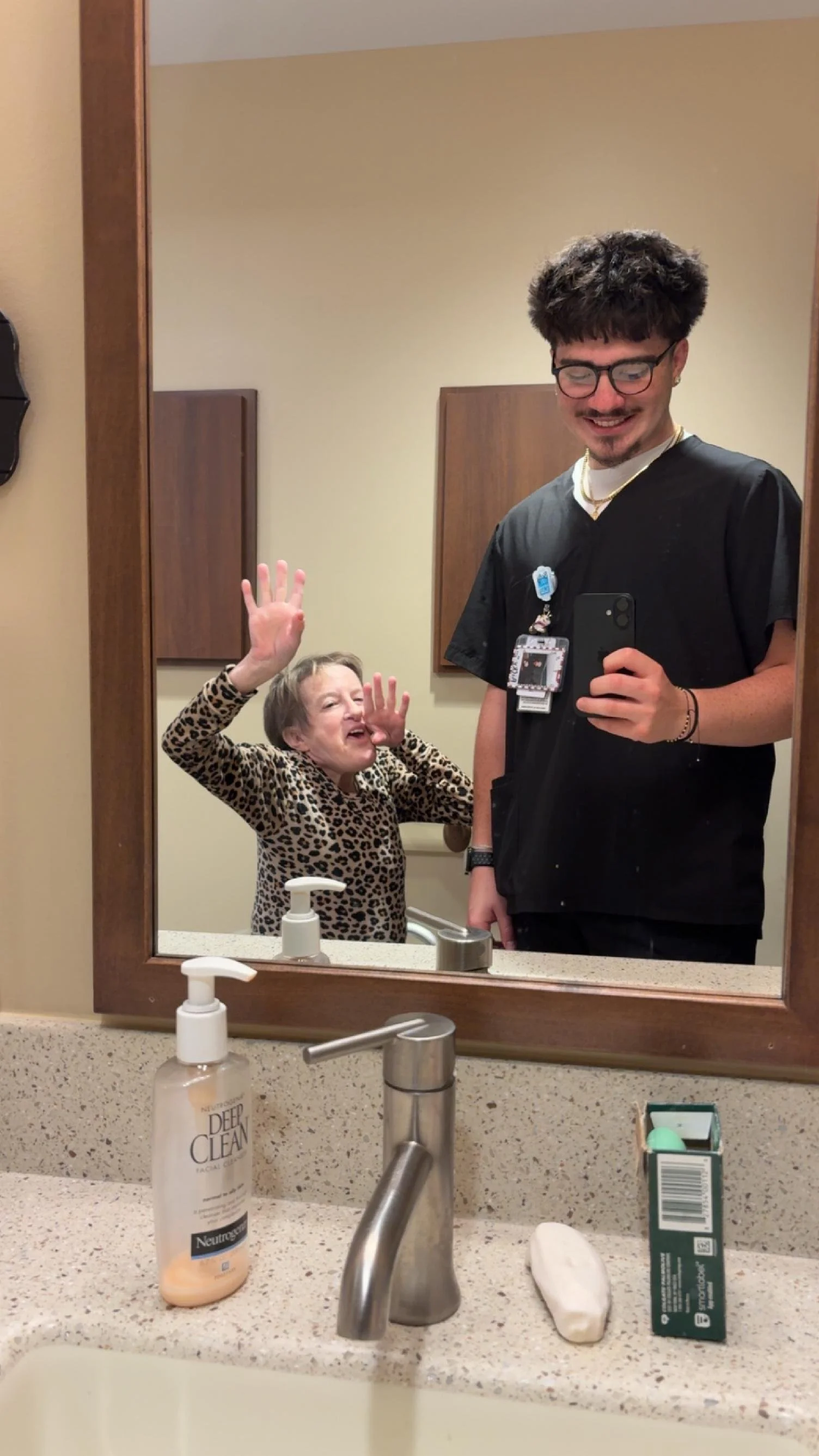 A young woman and a young man are taking a mirror selfie in a bathroom. The woman is smiling, waving, and wearing a leopard print top. The man is smiling, holding a phone, and wearing glasses and black scrubs. The bathroom has beige walls, wood accen