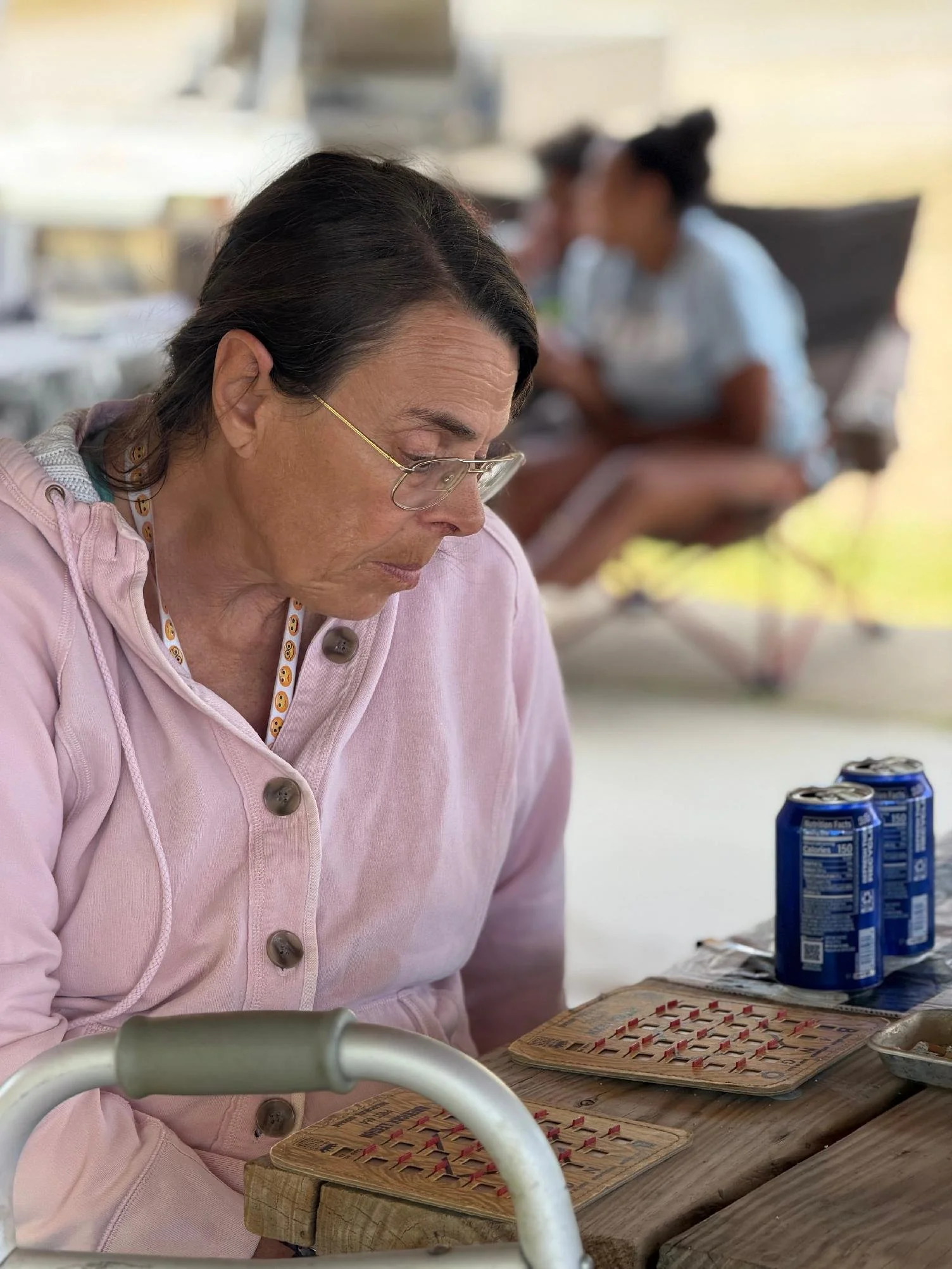 An older woman with glasses, wearing a pink shirt, playing bingo at a table with bingo cards and cans of soda in front of her, with two younger women in the background also playing.