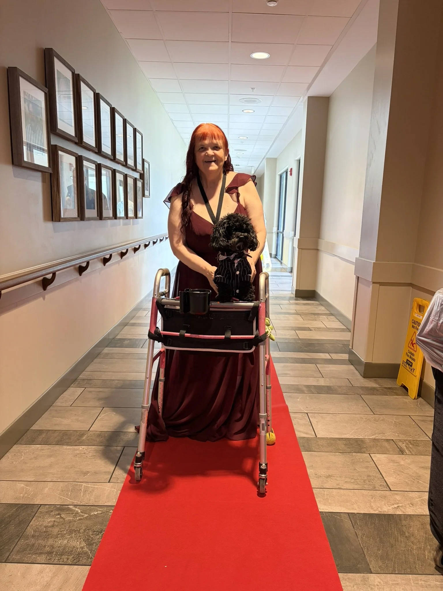 A woman with red hair and a maroon dress standing behind a walker on a red carpet in a corridor with framed pictures on the wall.
