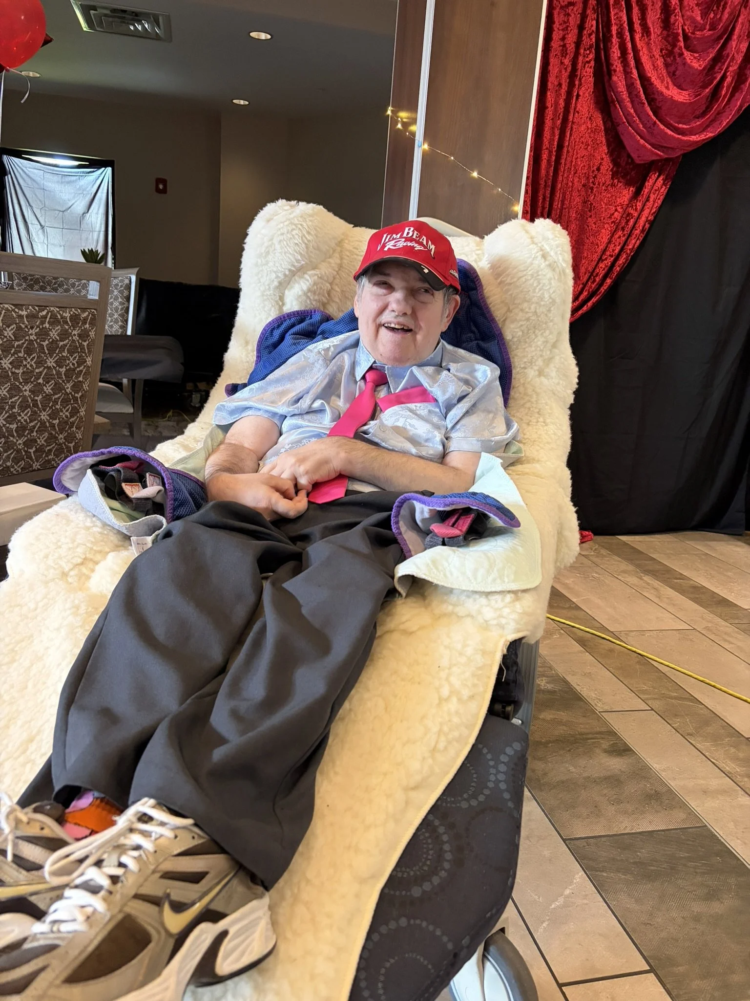 A man with gray hair smiling and dressed casually, wearing a red cap and a pink tie, seated comfortably on a plush chair with a cream-colored blanket. The background features red curtains and a string of small decorative lights.