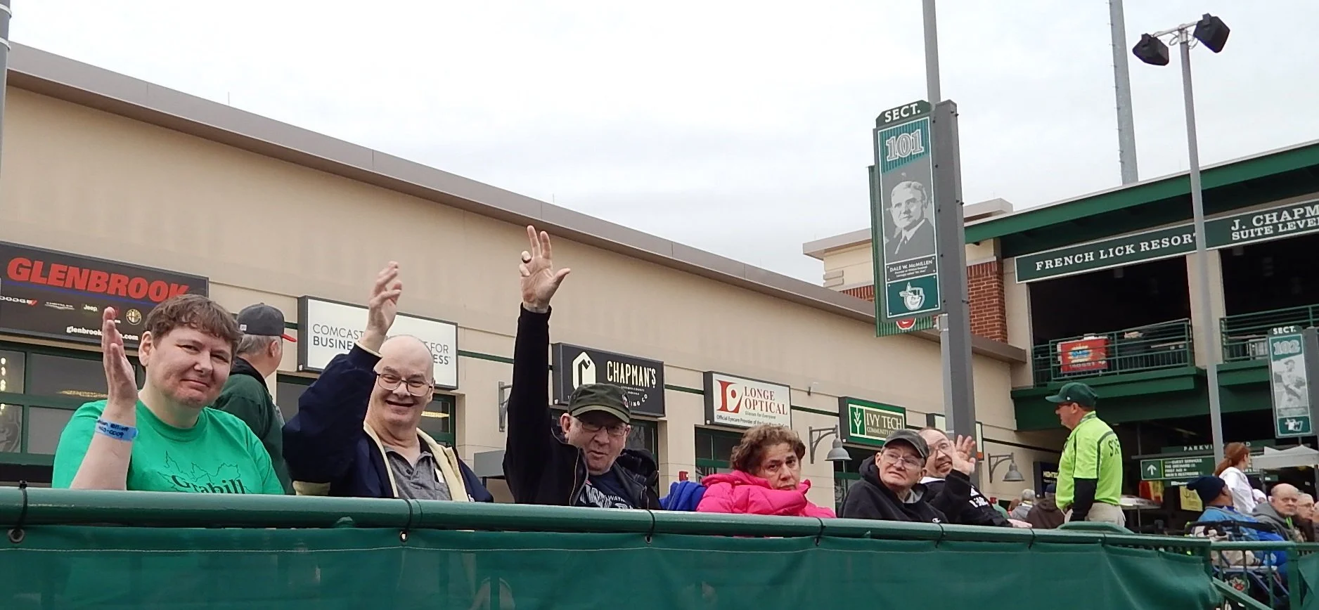 People sitting in a stadium or sports venue, waving and smiling, with signs and shops in the background.