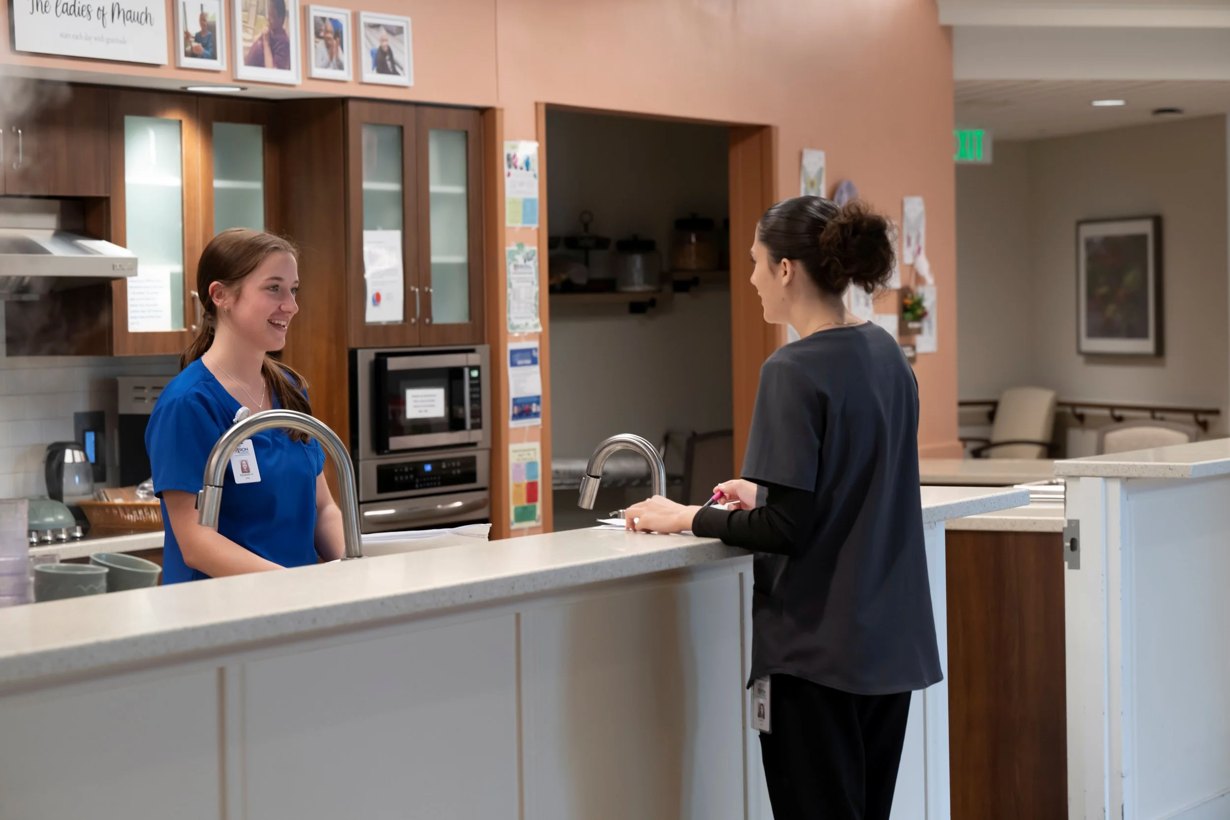 Healthcare worker in blue scrubs and a woman in black scrubs standing at a hospital or clinic reception desk, engaging in conversation. The background shows hospital kitchen appliances, framed photos on the wall, and an exit sign.