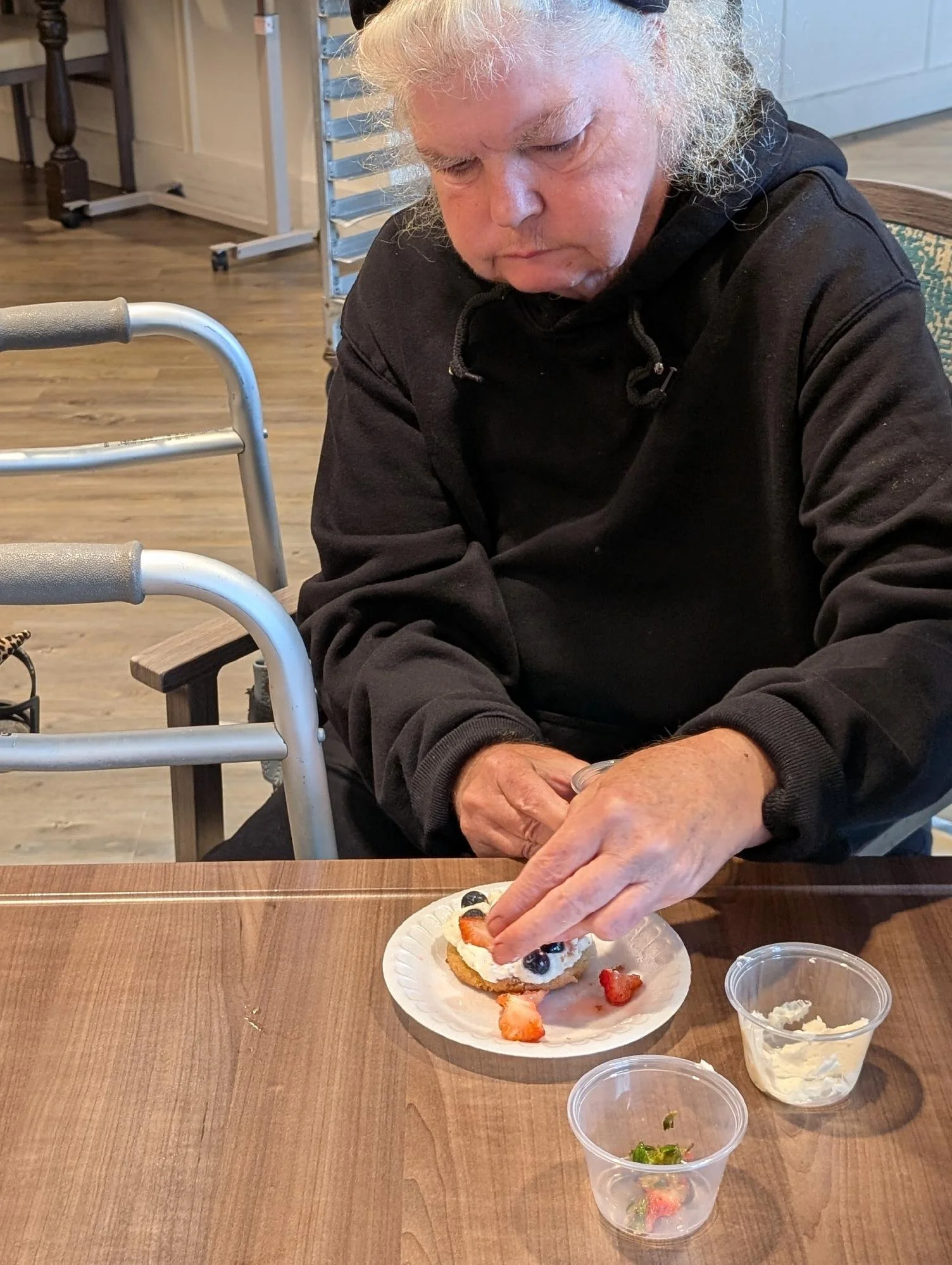 An elderly person with white hair and a black hoodie is sitting at a wooden table, preparing or eating a dessert with various berries on a paper plate. There are small cups with toppings or sides, and a walker is placed next to them.