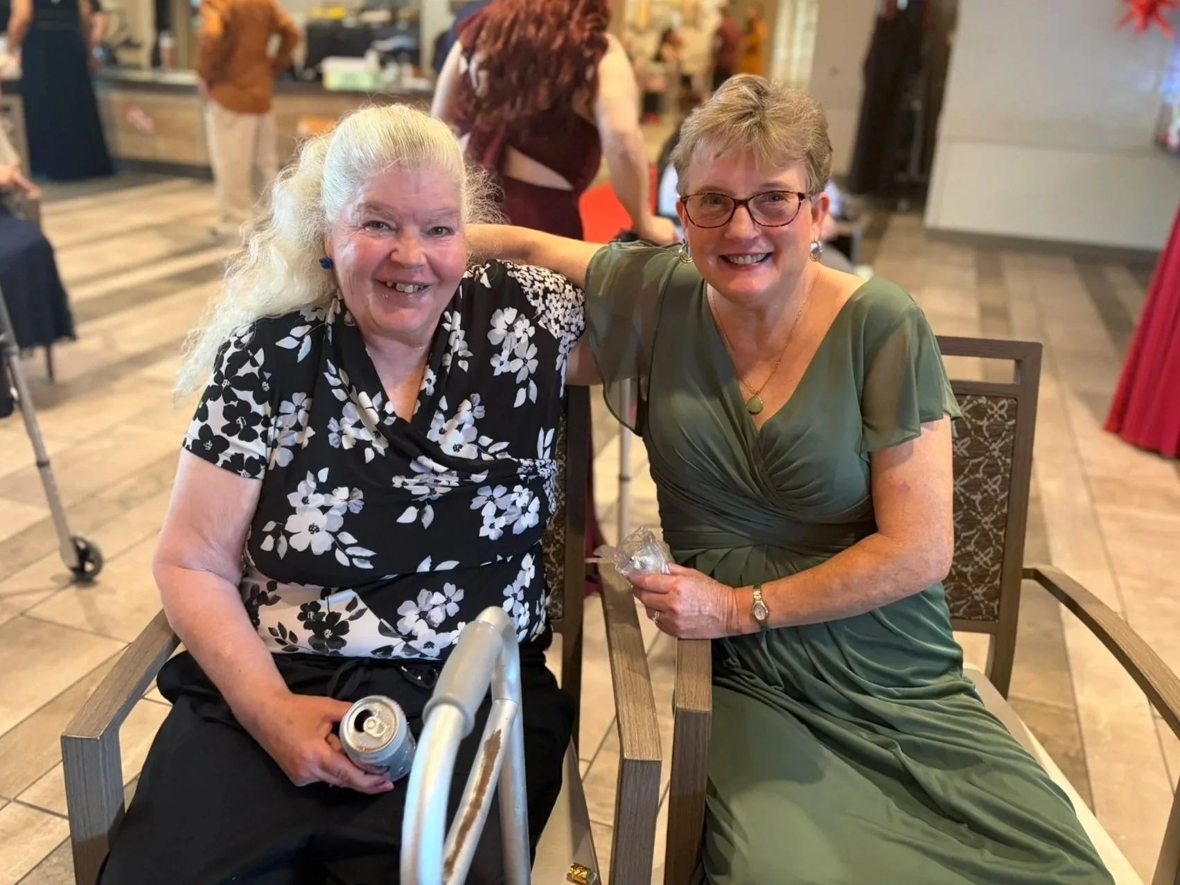 Two women sitting at a table in a restaurant or cafe, smiling at the camera. One woman has long, curly white hair and is wearing a black and white floral top. The other woman has short, gray hair and glasses, wearing a green dress, holding a wrapped gift or treat in her hand.