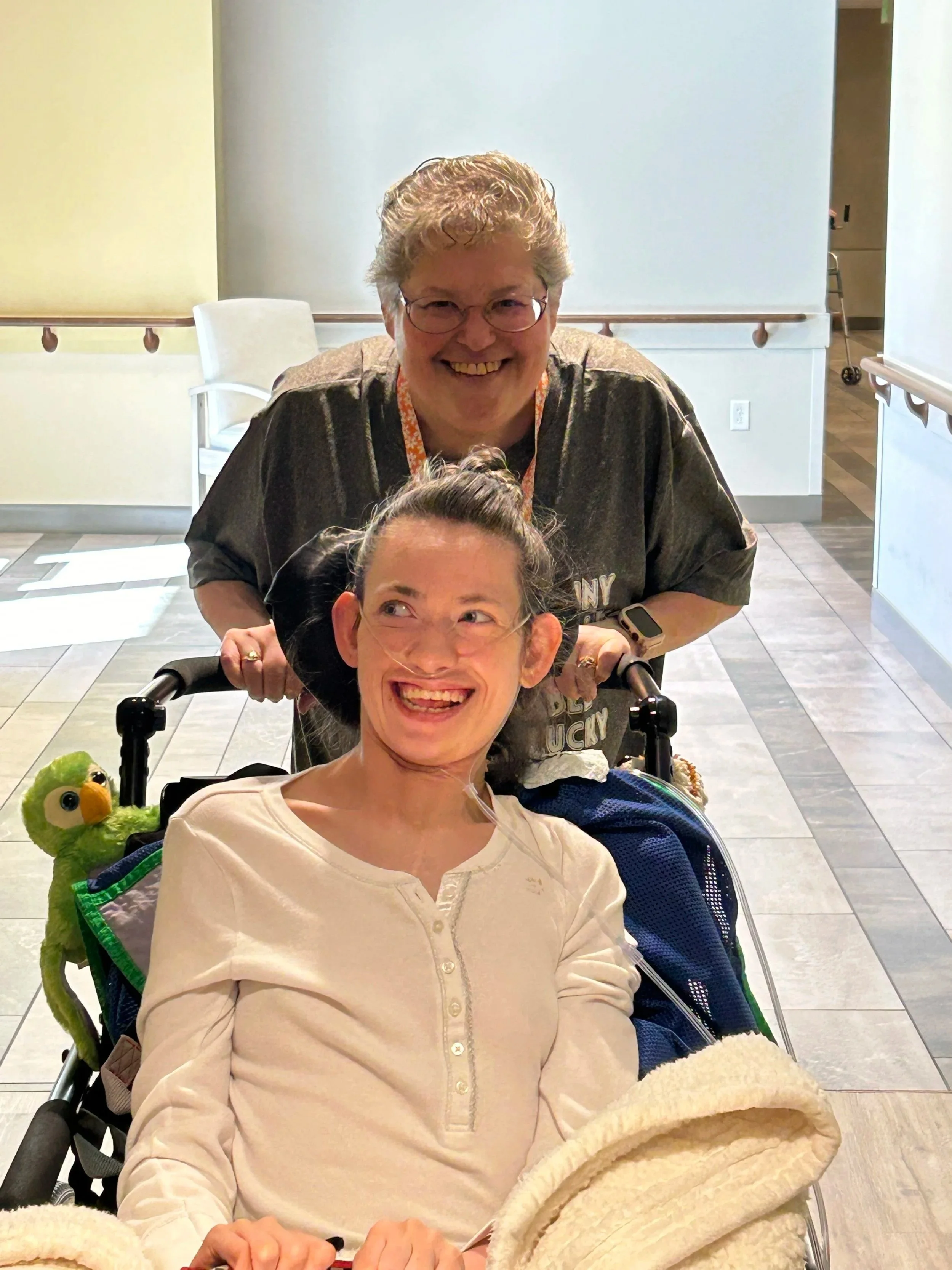 A man with glasses and curly hair standing behind a woman in a wheelchair in a hospital or care facility, both smiling. The woman has a nasal oxygen tube and is wearing a beige shirt, with a plush green toy bird on her chair. The background shows hospital or care facility furnishings.