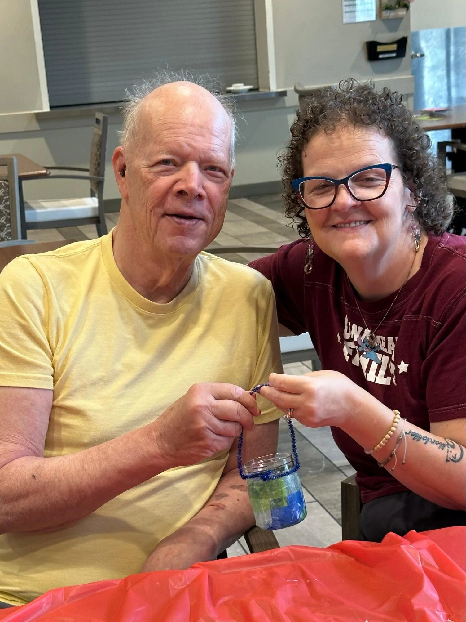 A smiling elderly man in a yellow t-shirt and a woman with glasses and curly hair in a maroon t-shirt sit at a table. They are holding a beaded bracelet together, and there is a jar with a layered drink in front of them. The background shows a restau